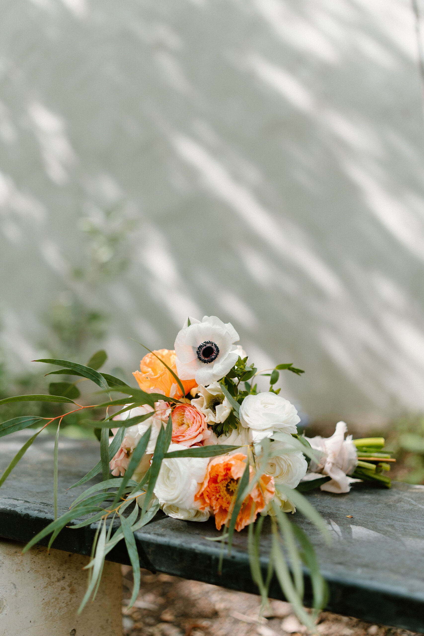 bridal bouquet of greenery, poppies, eucalyptus, and other orange flowers, positioned on a bench outdoors against a white wall with dappled shadows during a spring wedding at the Maas Building