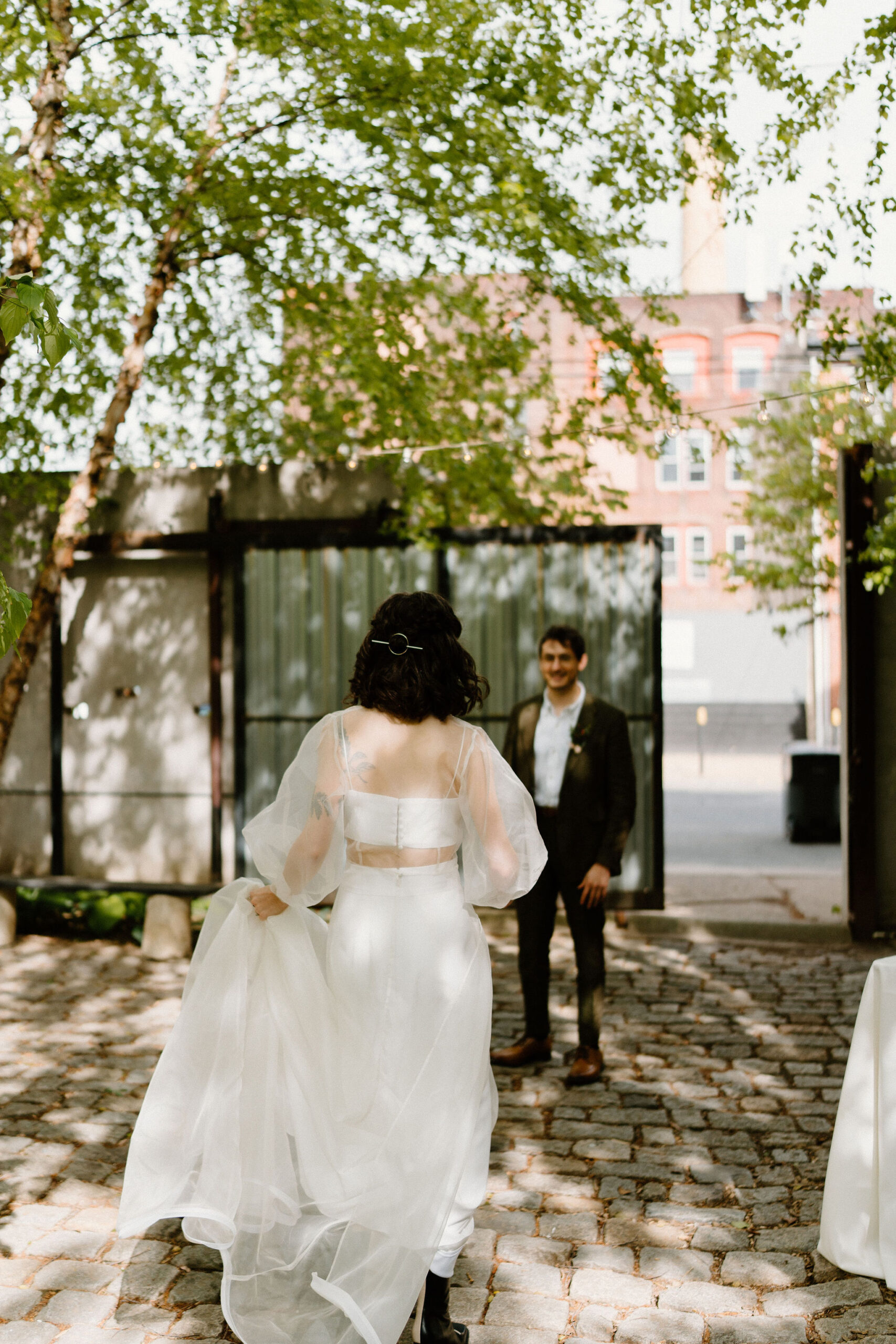 bride (brown hair, sheer cape, two piece gown) walking up to her groom (brown hair, green suit) in a lush green cobblestone courtyard at the Maas Build