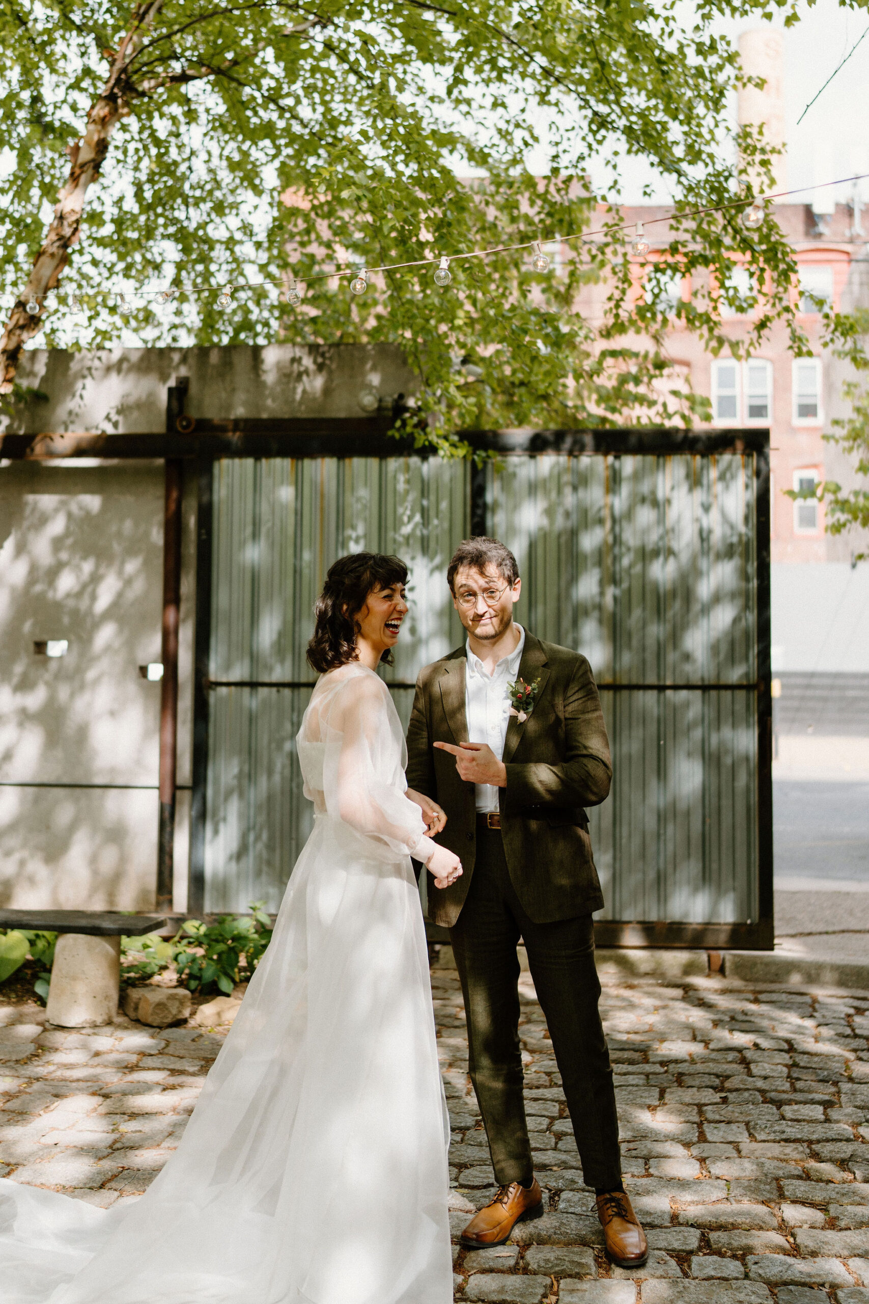 bride (brown hair, sheer cape, two piece gown) and groom (brown hair, green suit) in a lush green cobblestone courtyard at the Maas Build, her laughing at him and him smirking at the camera, pointing at her