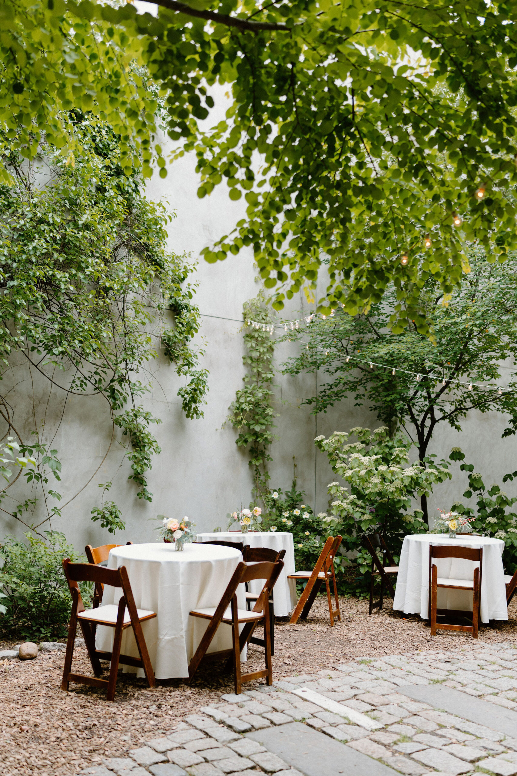 wide image of three tables set up with wooden chairs, white tablecloths, and a small bouquet of flowers, inside a lush springtime courtyard garden with cobblestone