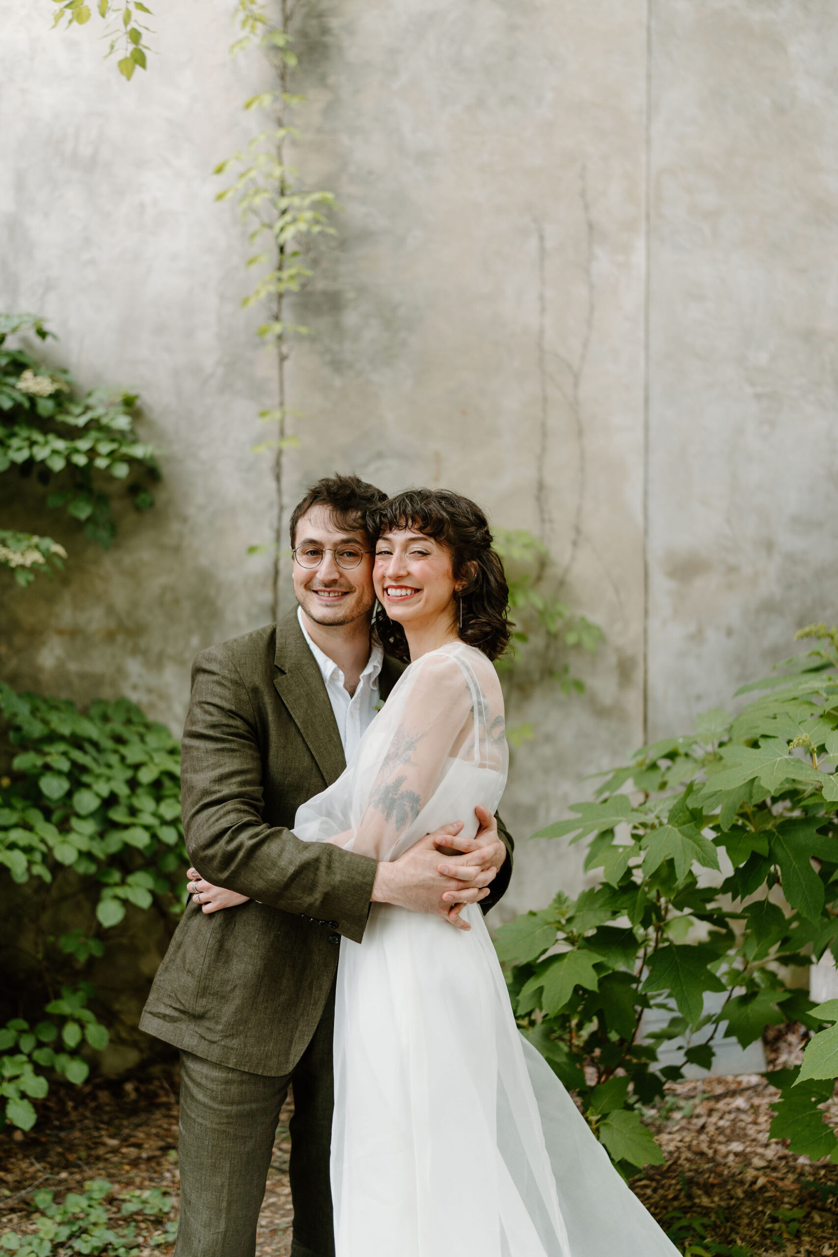 bride and groom embracing and smiling at the camera in a lush green cobblestone courtyard before their spring wedding at the Maas Building