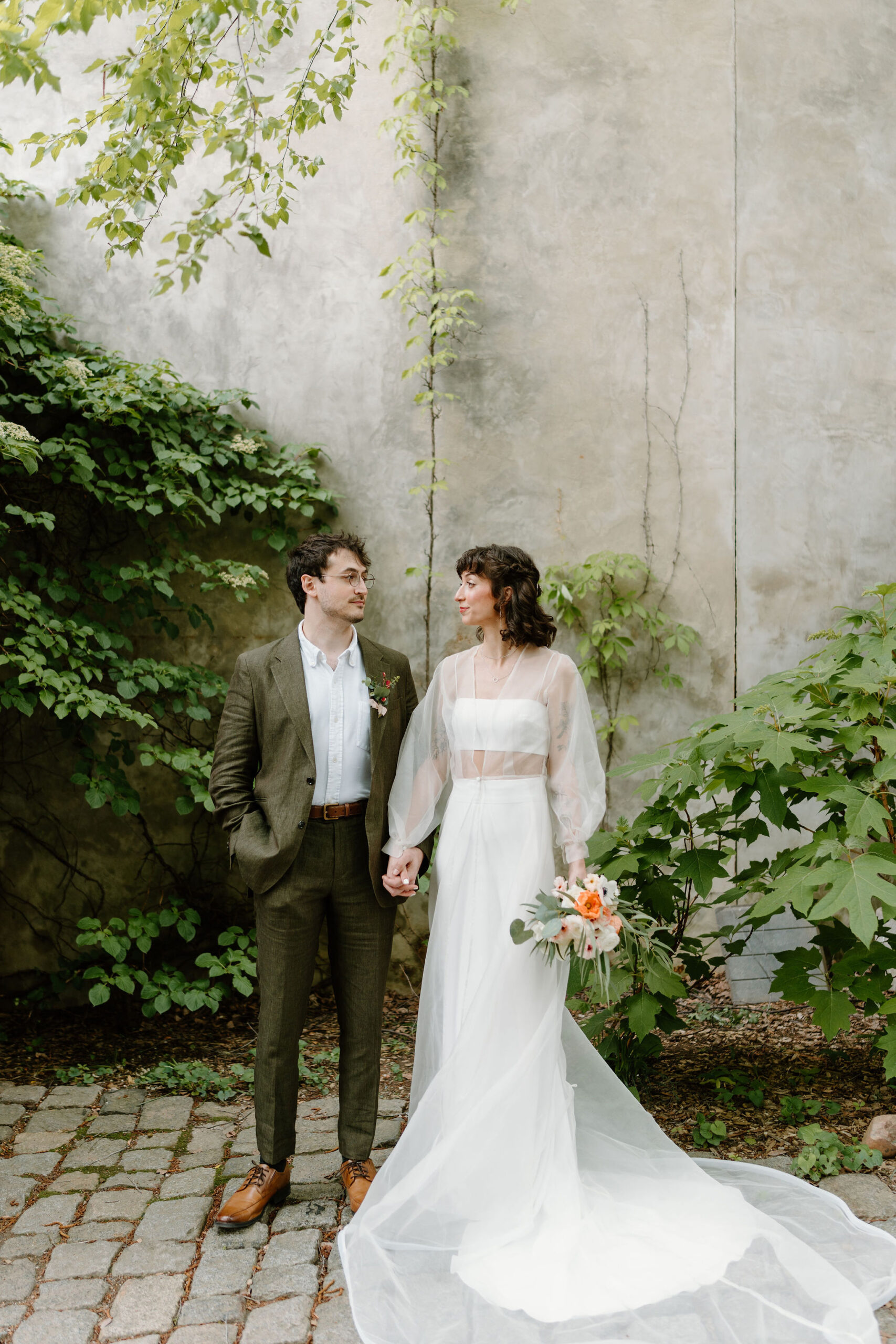 bride (two piece cropped dress with a sheer long sleeve cape, curly short brown hair with bangs) holding her bouquet at her side and holding groom's hand (brown hair, olive linen suit, brown shoes), smiling softly at each other in a lush green cobblestone courtyard at the Maas Building