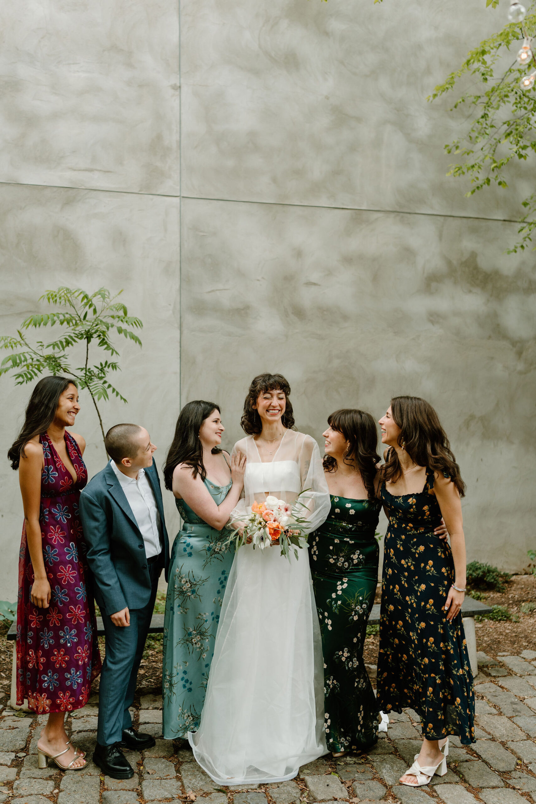 bride (wearing white) and five of her friends (in various colorful and eclectic dresses and a blue suit) smiling at each other in a lush green cobblestone courtyard for a spring wedding