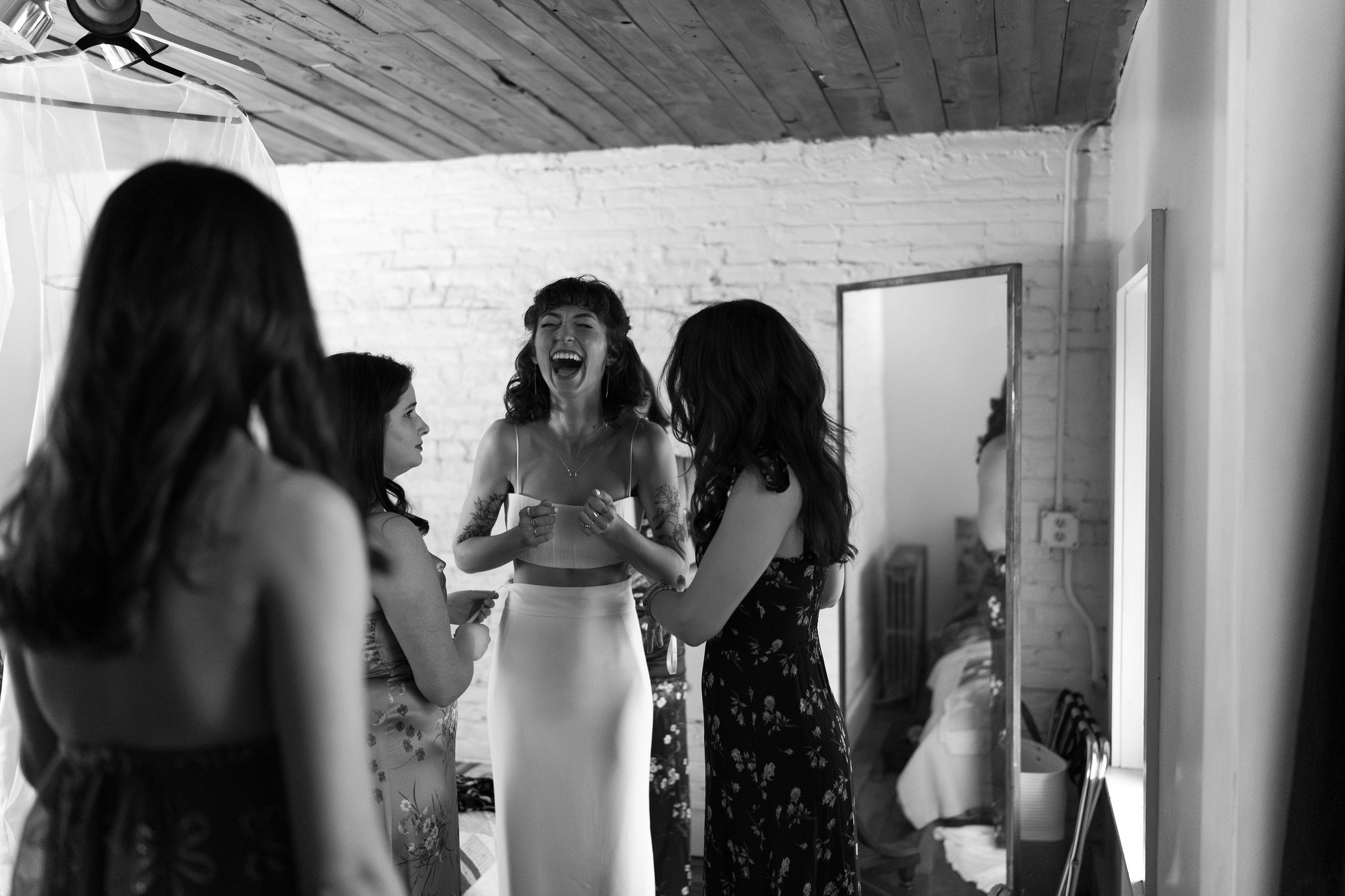 black and white image of a bride in a two piece wedding dress laughing, while four of her friends help her get into the dress at the Maas Building