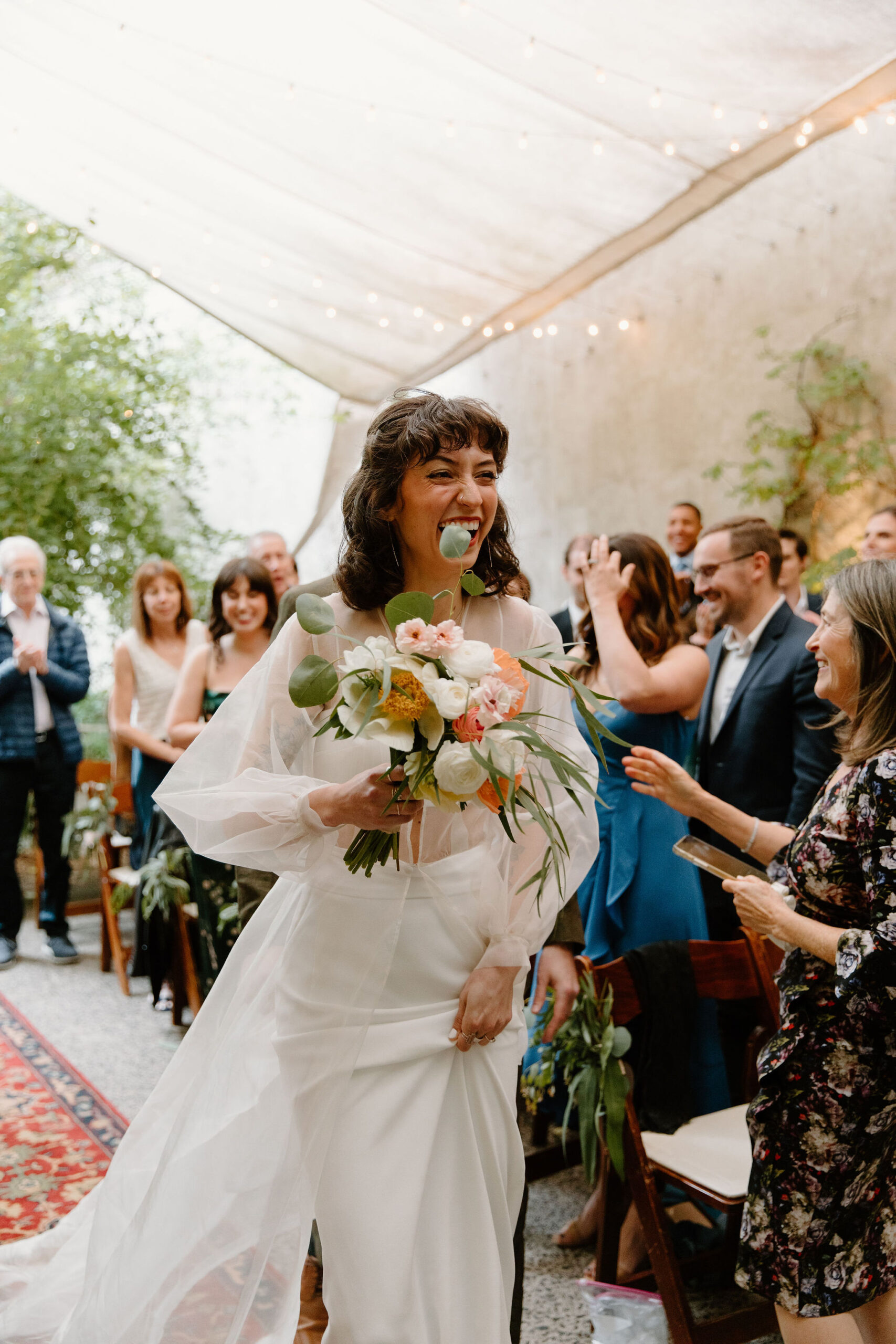 bride walking down the aisle to her spring wedding ceremony at the Maas building, holding her flowers and laughing while her guests smile behind her