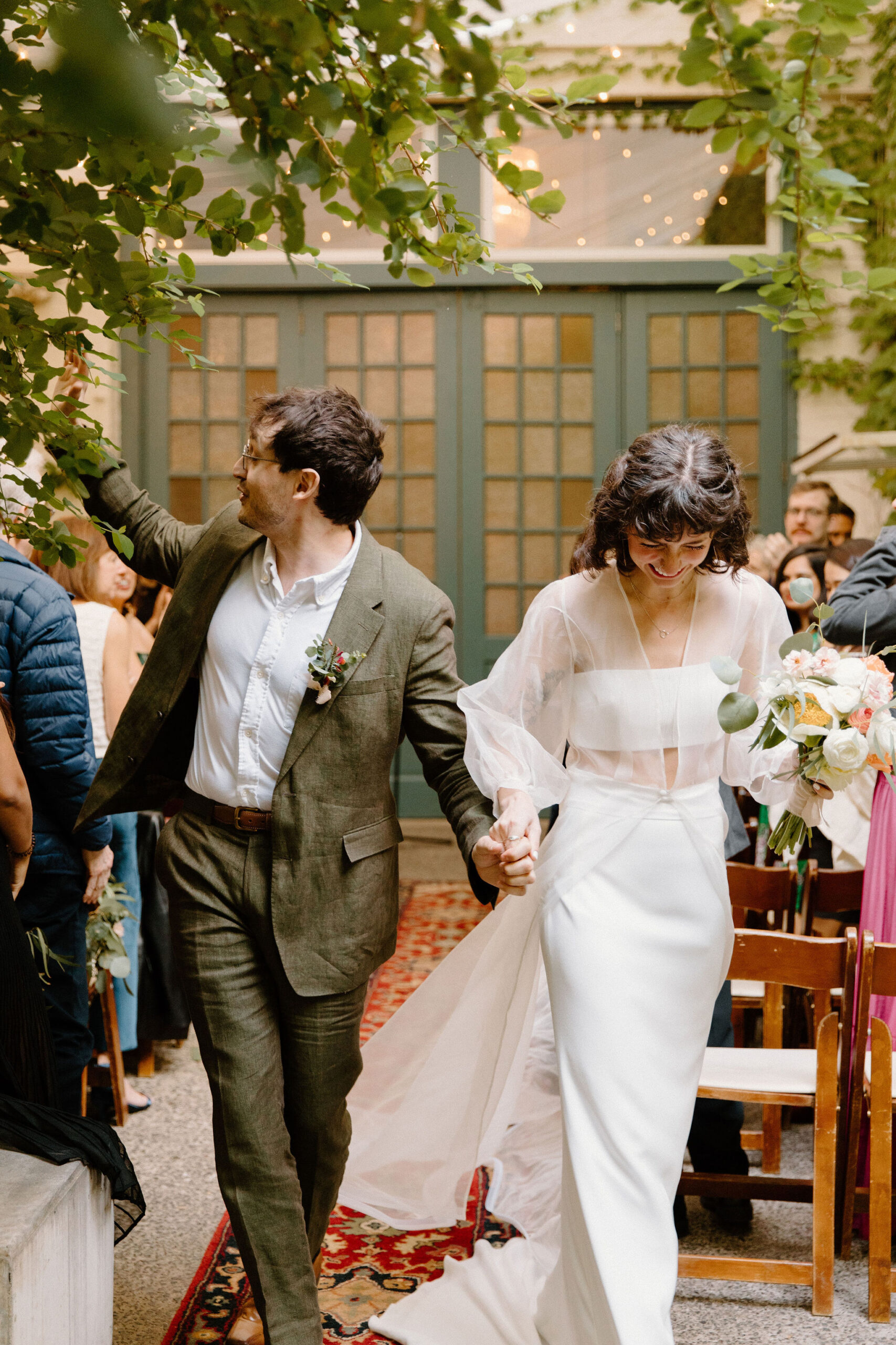 bride and groom holding hands as they exit their spring wedding ceremony at the Maas Building, bride smiling down while groom high fives a guest