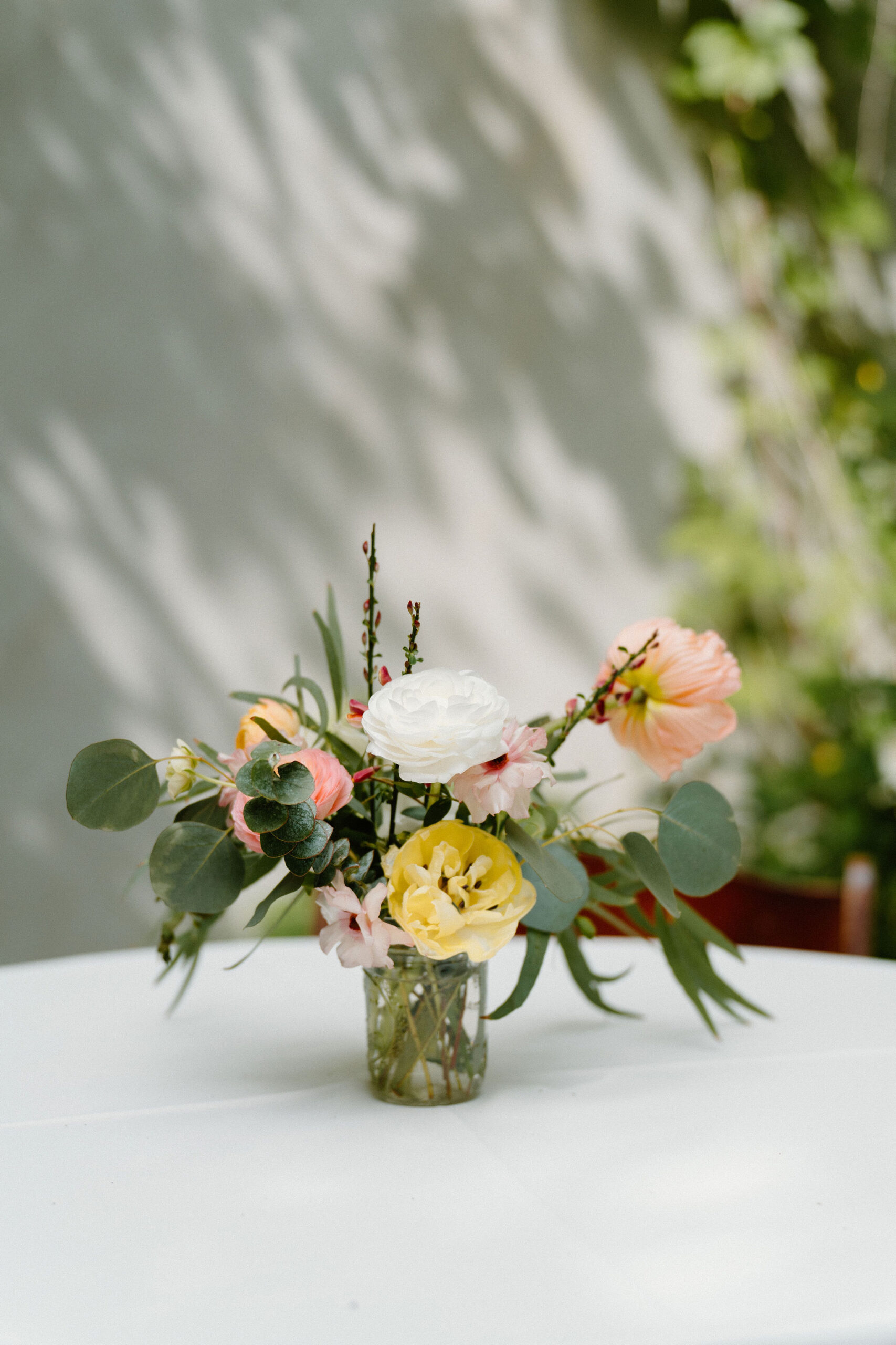 a small centerpiece flower vase holding poppies, peonies, and eucalyptus on a white tablecloth, outdoors against a white wall with lots of midday shadows across it during a spring wedding at the Maas Building