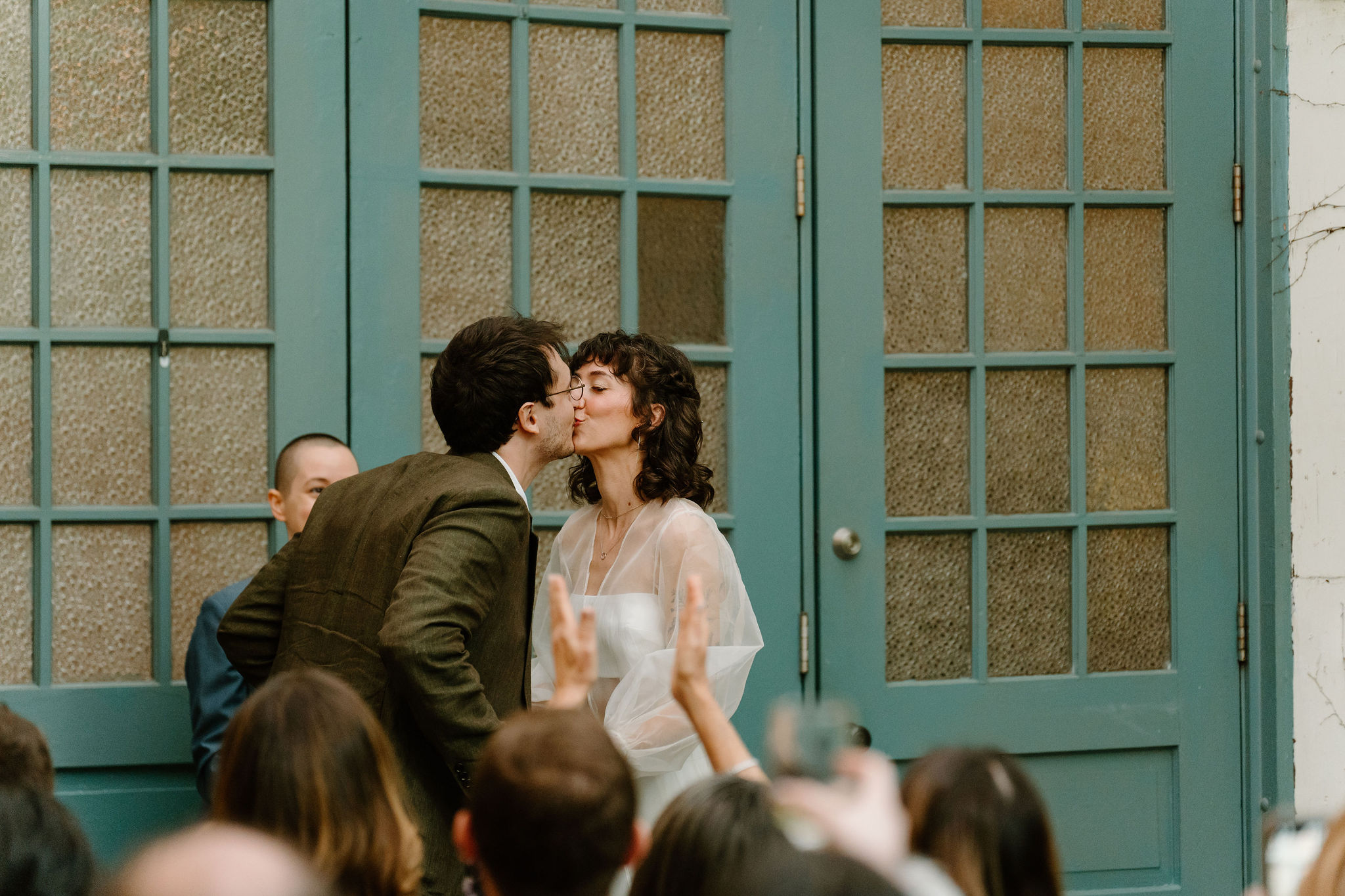 bride and groom exchanging their first kiss during their spring wedding ceremony at the Maas Building, in front of large teal doors while their guests watch