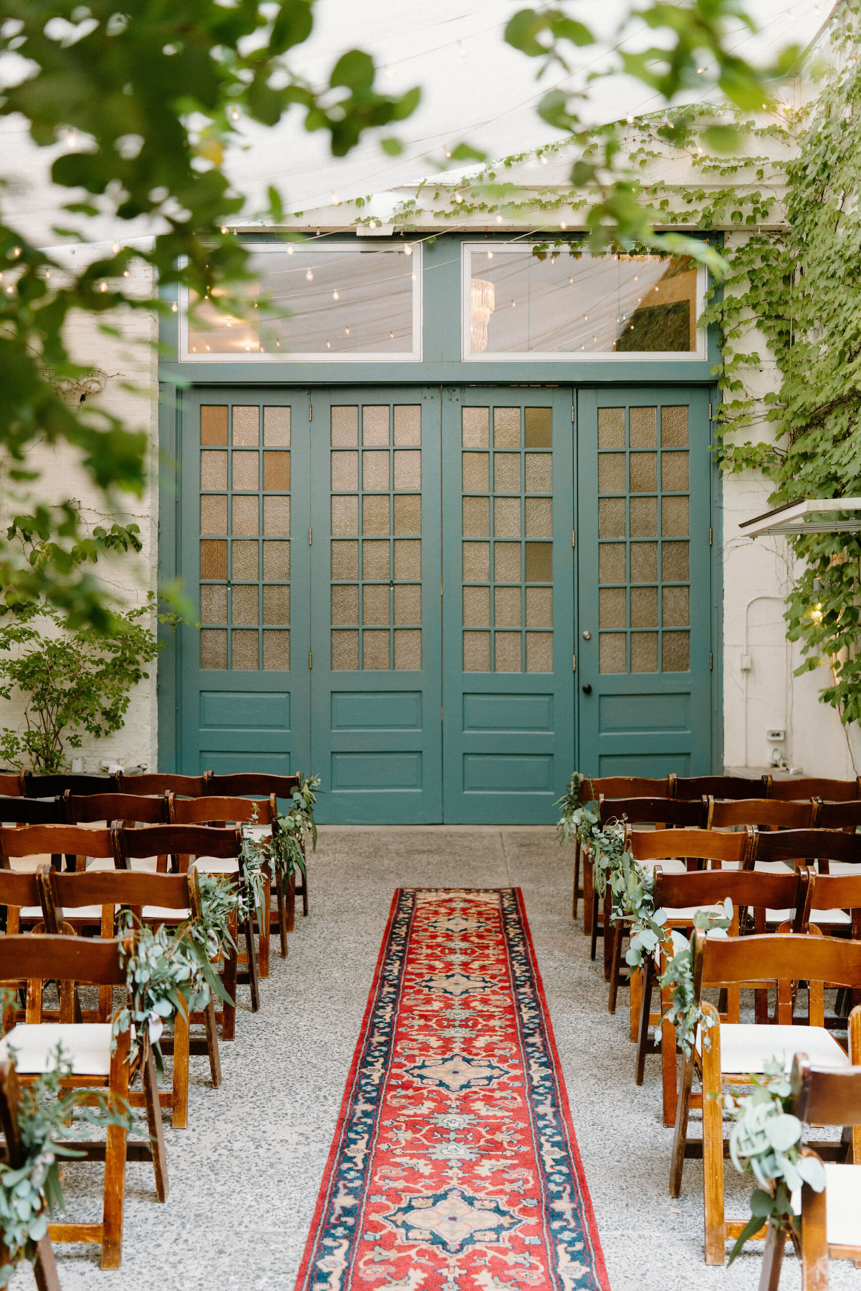 an outdoor courtyard ceremony space at the Maas Building, against large teal doors with large windows, with two sets of wooden chairs around a center aisle with an antique style red carpet runner. There are vines and greenery growing all around the walls.