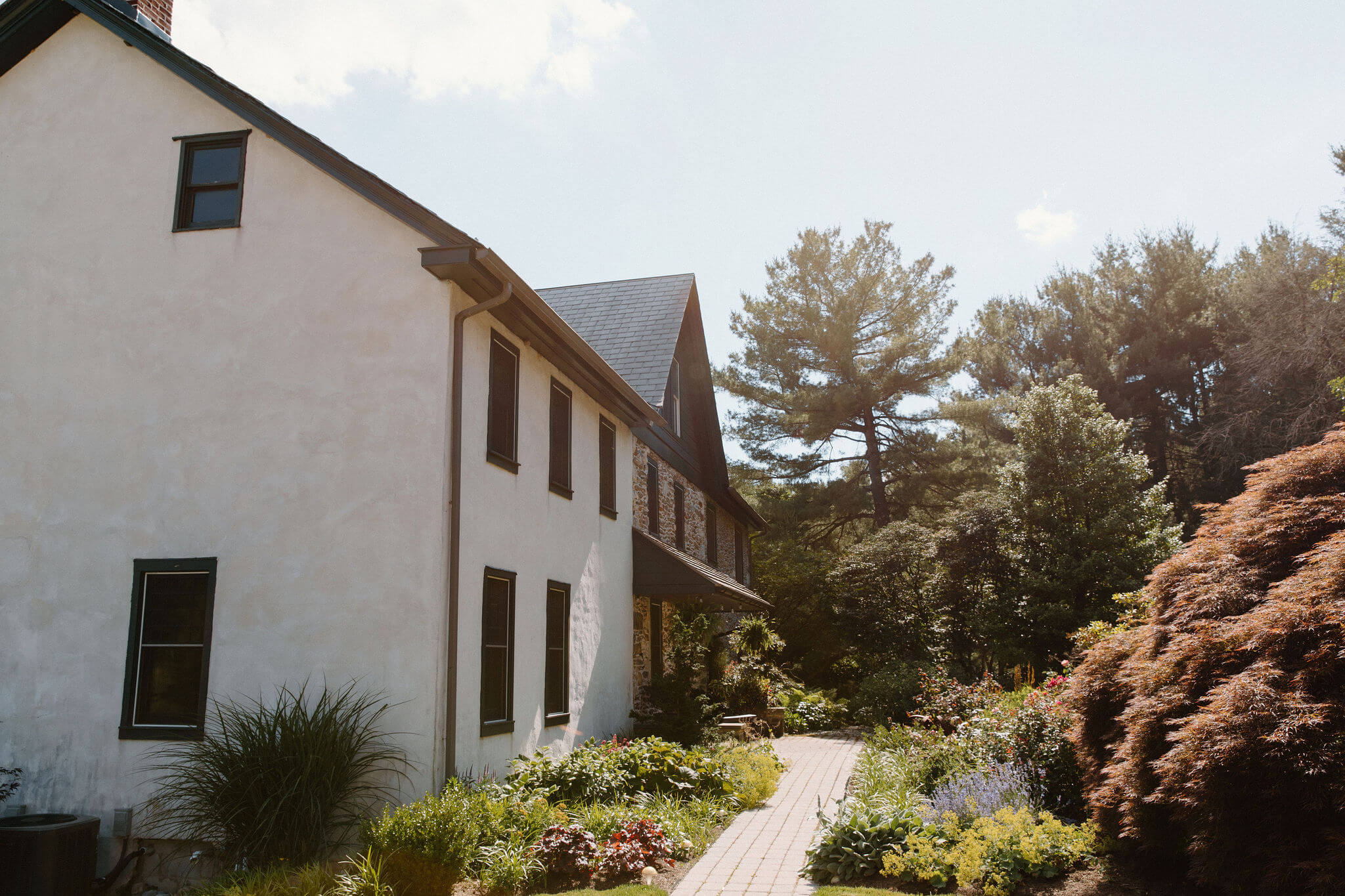a brick path cutting through a front garden full of lush greenery and flowering bushes, leading up to the front door of a stone farm house in Pennsylvania  with a stucco addition
