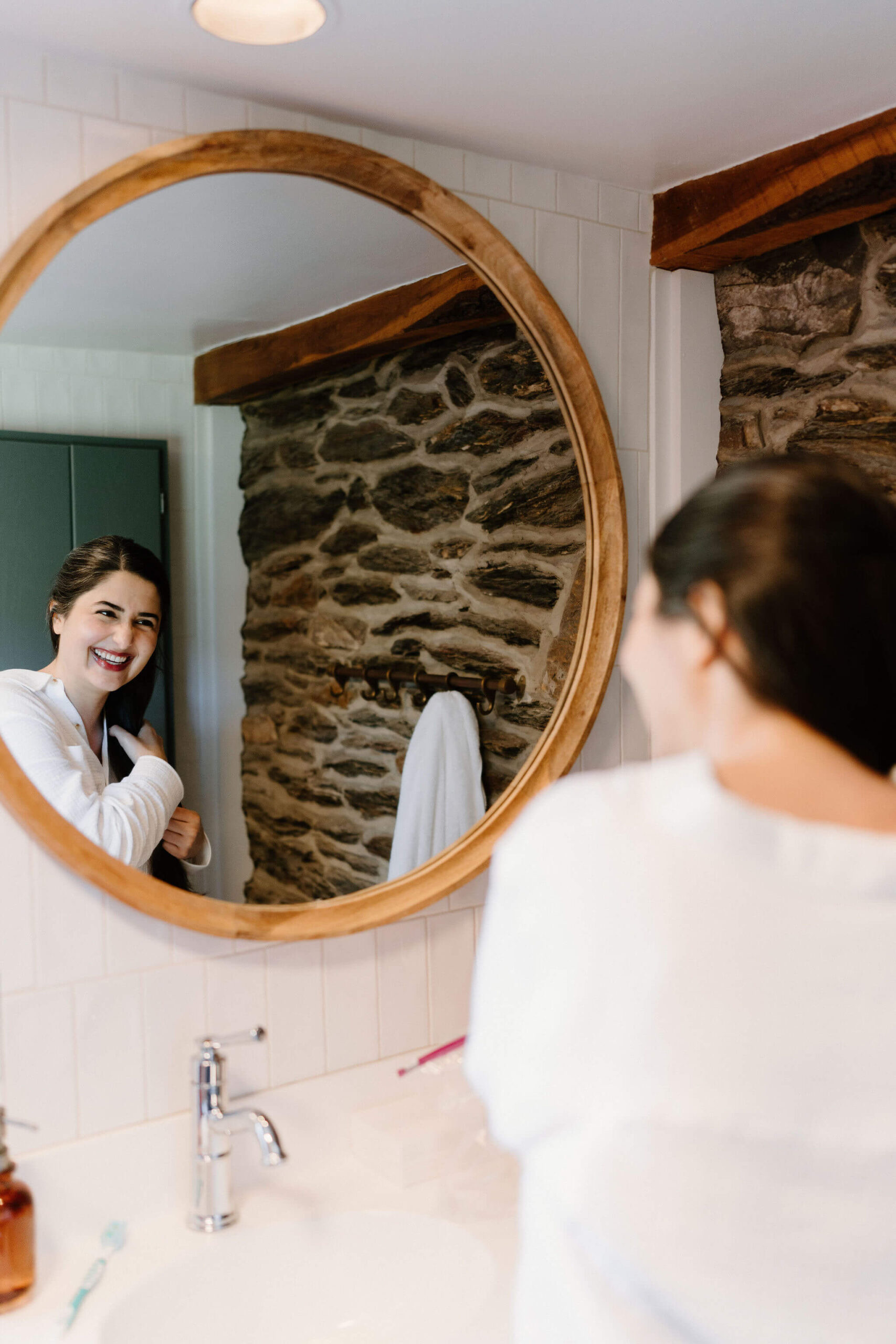bride smiling at herself in the bathroom mirror, while styling her long brown hair, getting ready for her elopement