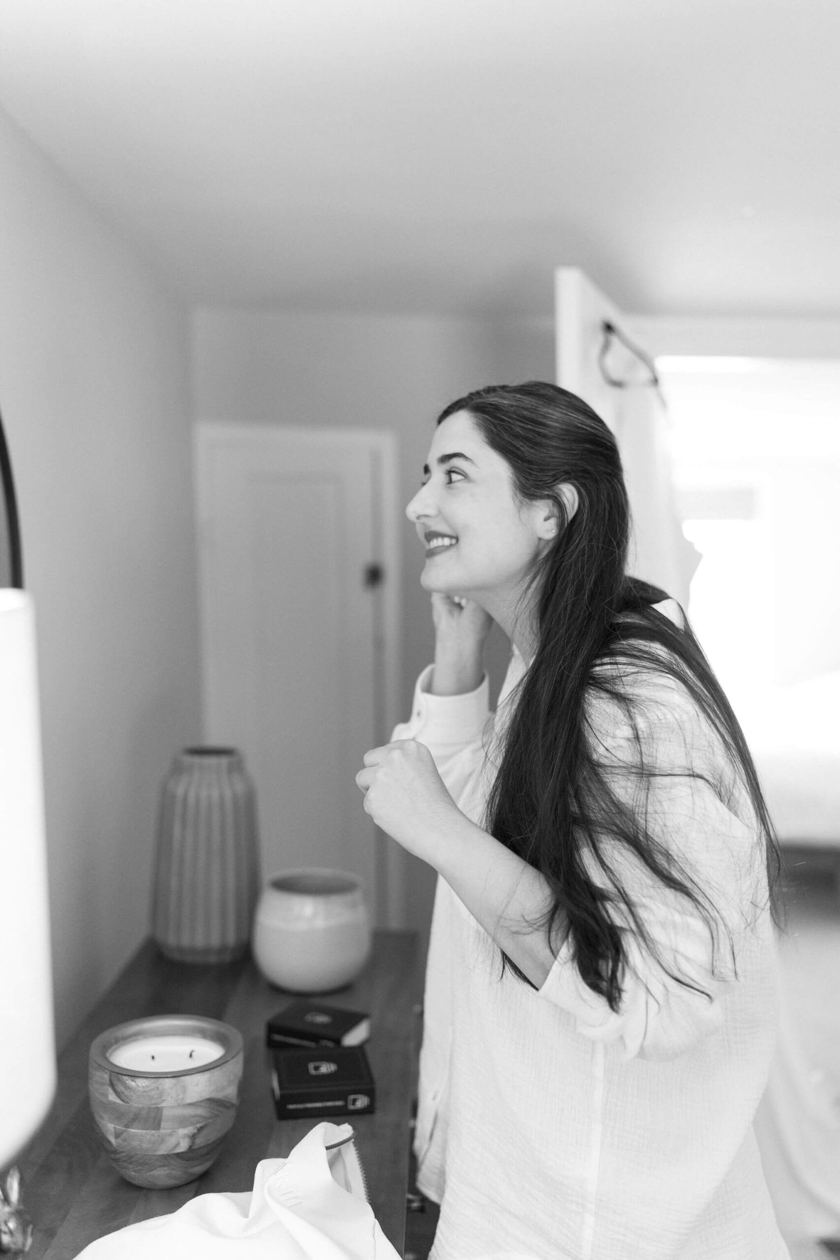 black and white image of a woman (bride, long brown hair) in a white button down shirt, looking in a bedroom mirror to put her earrings in and getting ready to elope