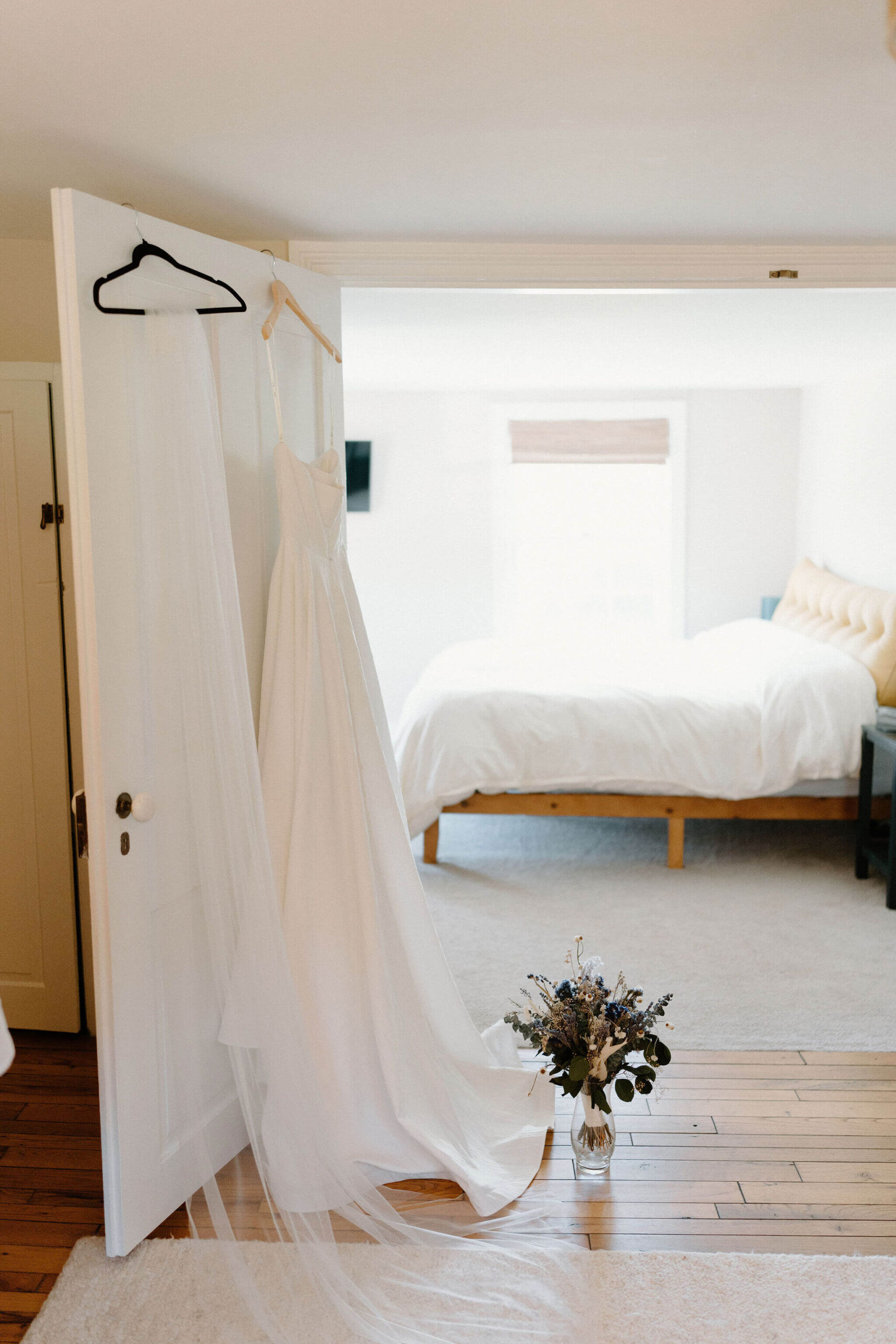 elopement dress and veil on hangers, hanging on a bedroom door. there is a vase of dried florals set on the floor next to the dress, and a made bed in the background.