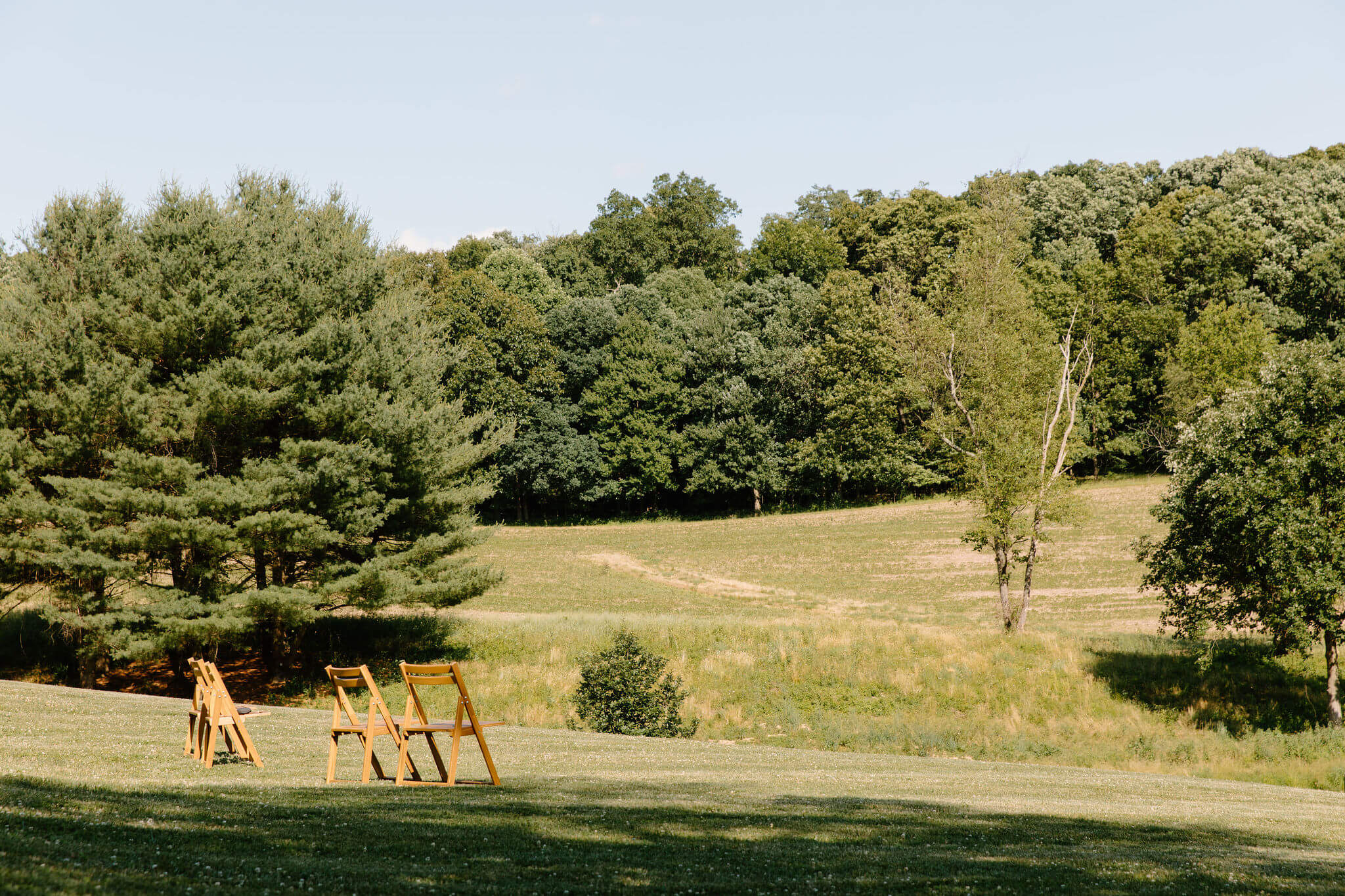 four wooden chairs placed in an open field for a farmhouse elopement in pennsylvania, with a rolling hill and forest in the background