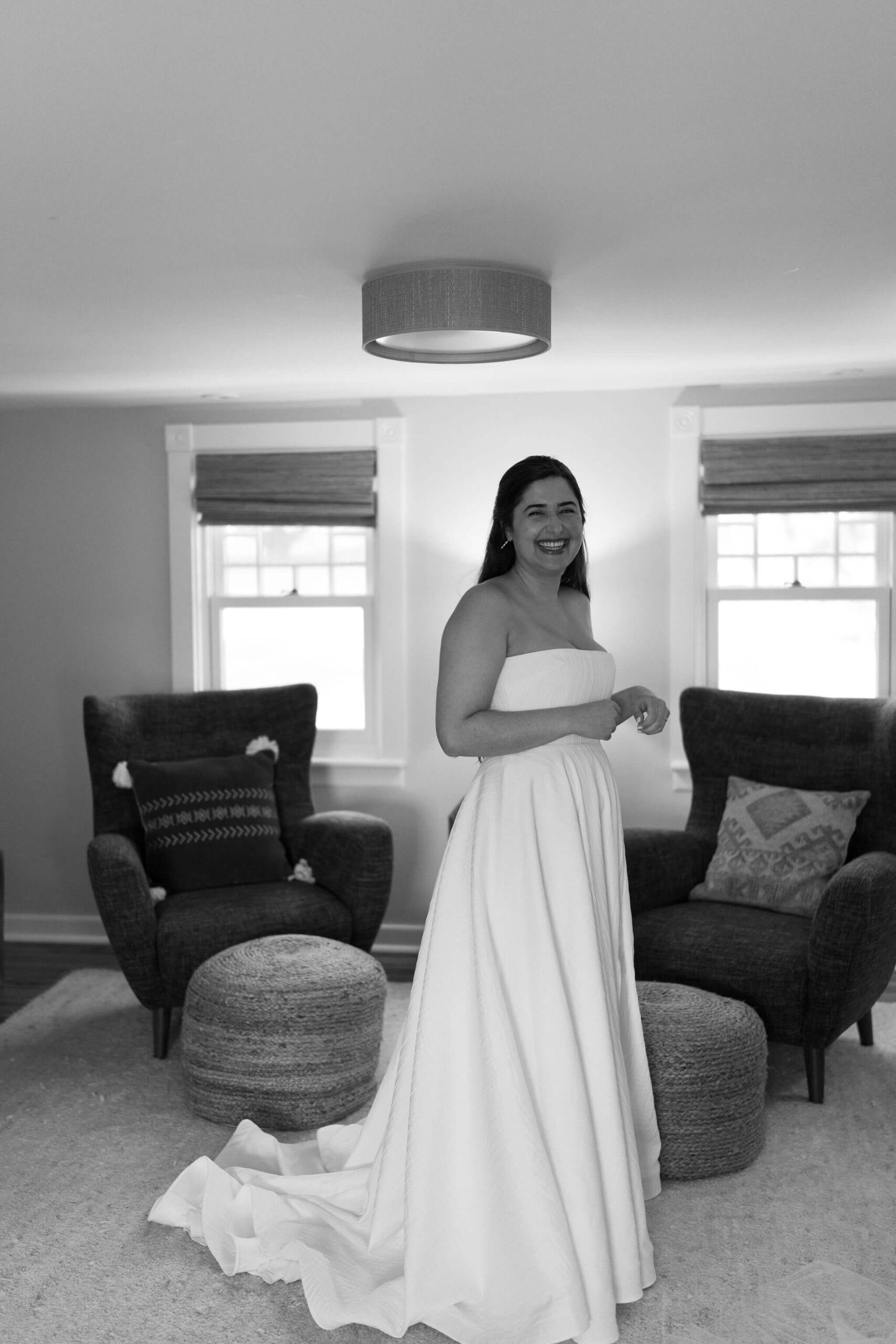 black and white image of a bride (long brown hair, strapless white wedding gown) standing in a farmhouse bedroom laughing toward the camera
