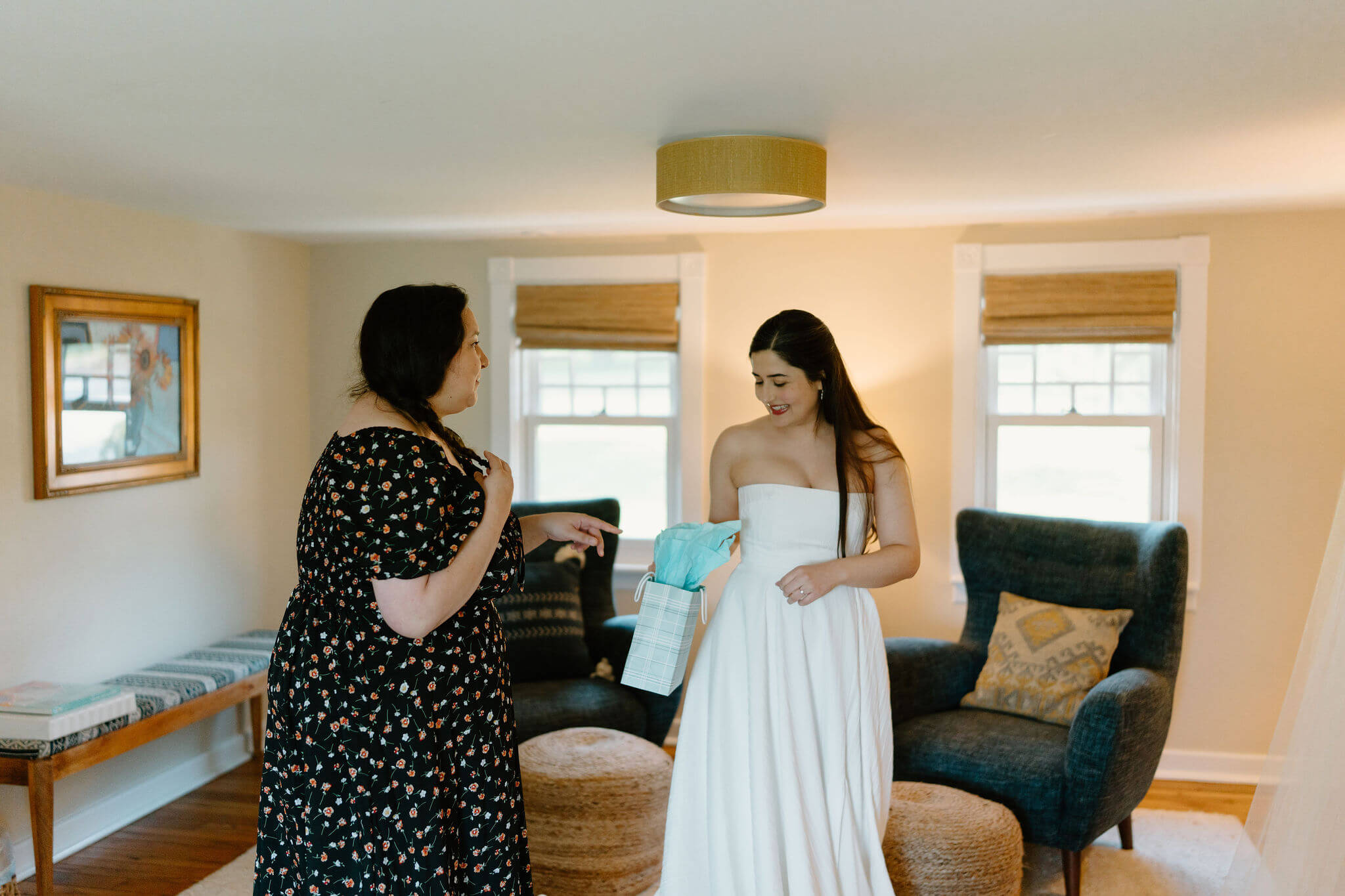 bride's friend in a black floral dress, giving the bride (long dark hair, white strapless dress) a gift before her elopement ceremony