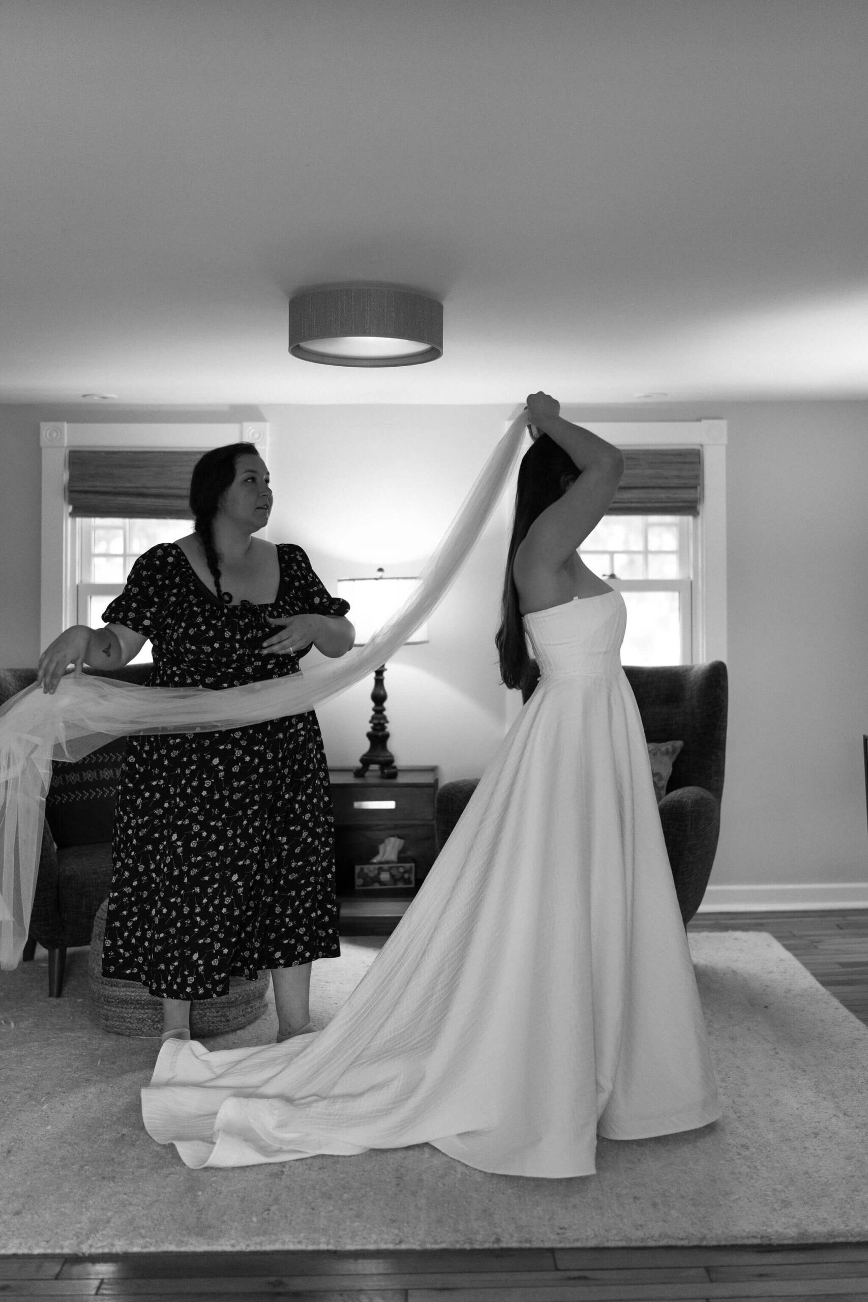 black and white image of bride's friend (black dress with floral accents) holding the end of a wedding veil while bride places it in her hair, wearing a long white bridal gown before her pennsylvania elopement