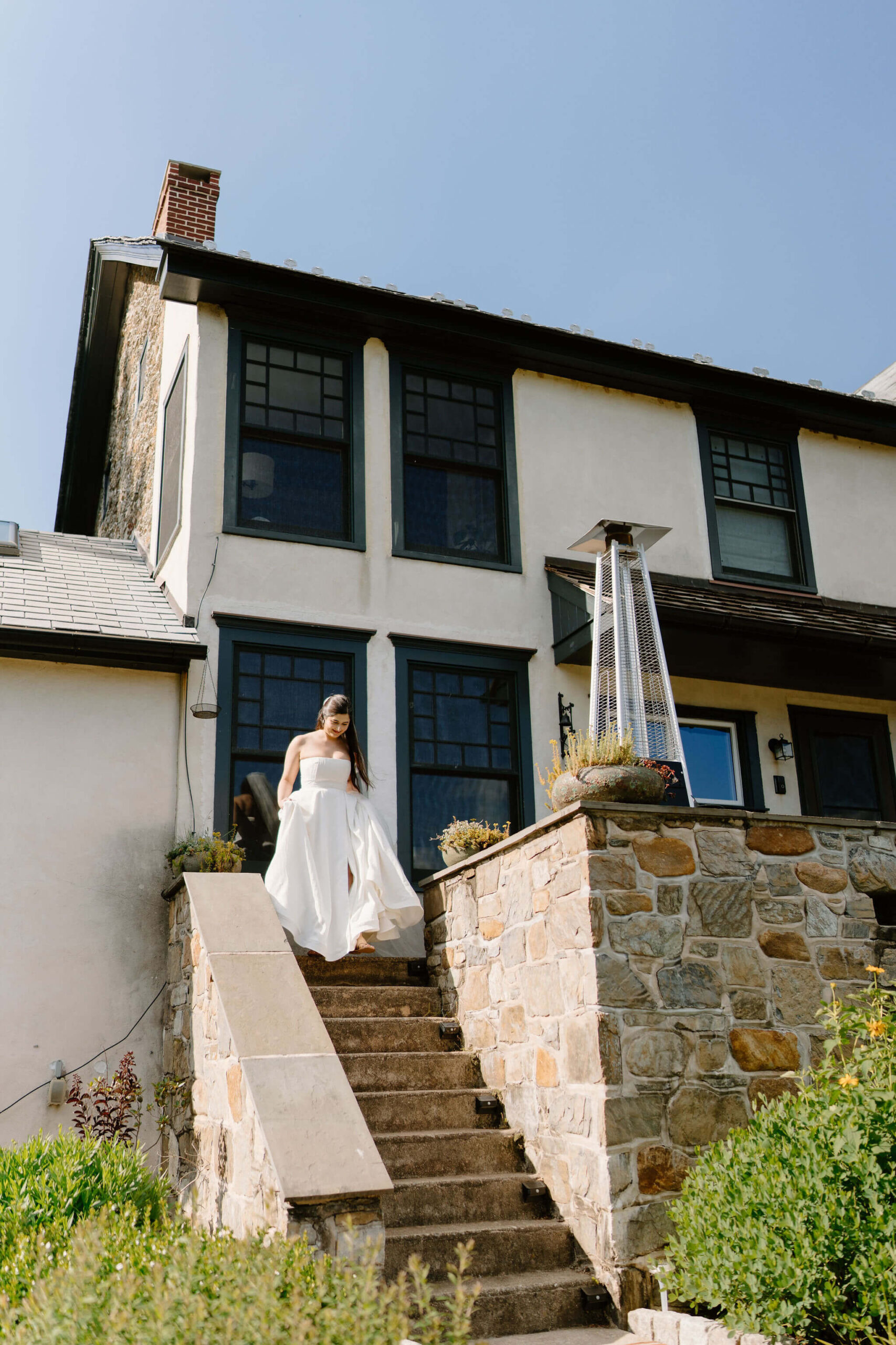 bride with brown hair walking down the stone stairs of a large pennsylvania farmhouse on a sunny day, holding the hem up of her strapless white wedding gown