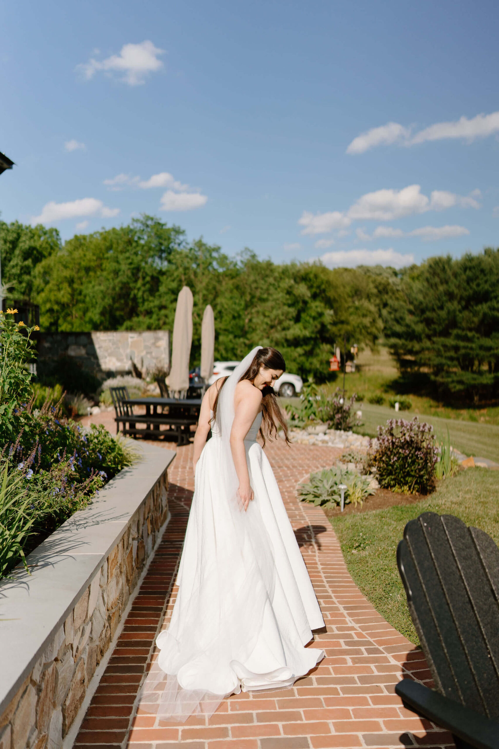 bride walking down a brick patio next to a stone wall, reaching behind her to straighten the train of her gown behind her, on the way to her elopement ceremony in pennsylvania