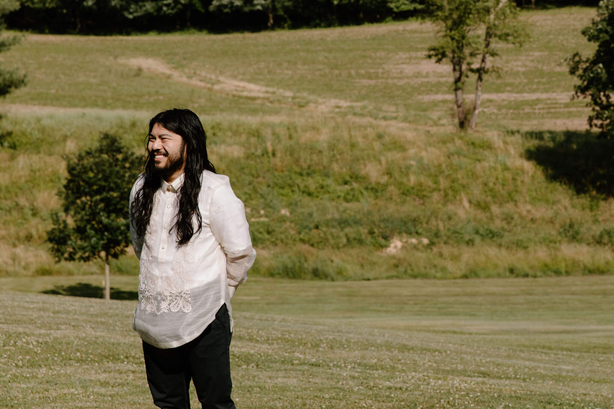 groom with long brown hair and a short beard, smiling off camera toward the bride, waiting for her to arrive to their elopement ceremony in a field