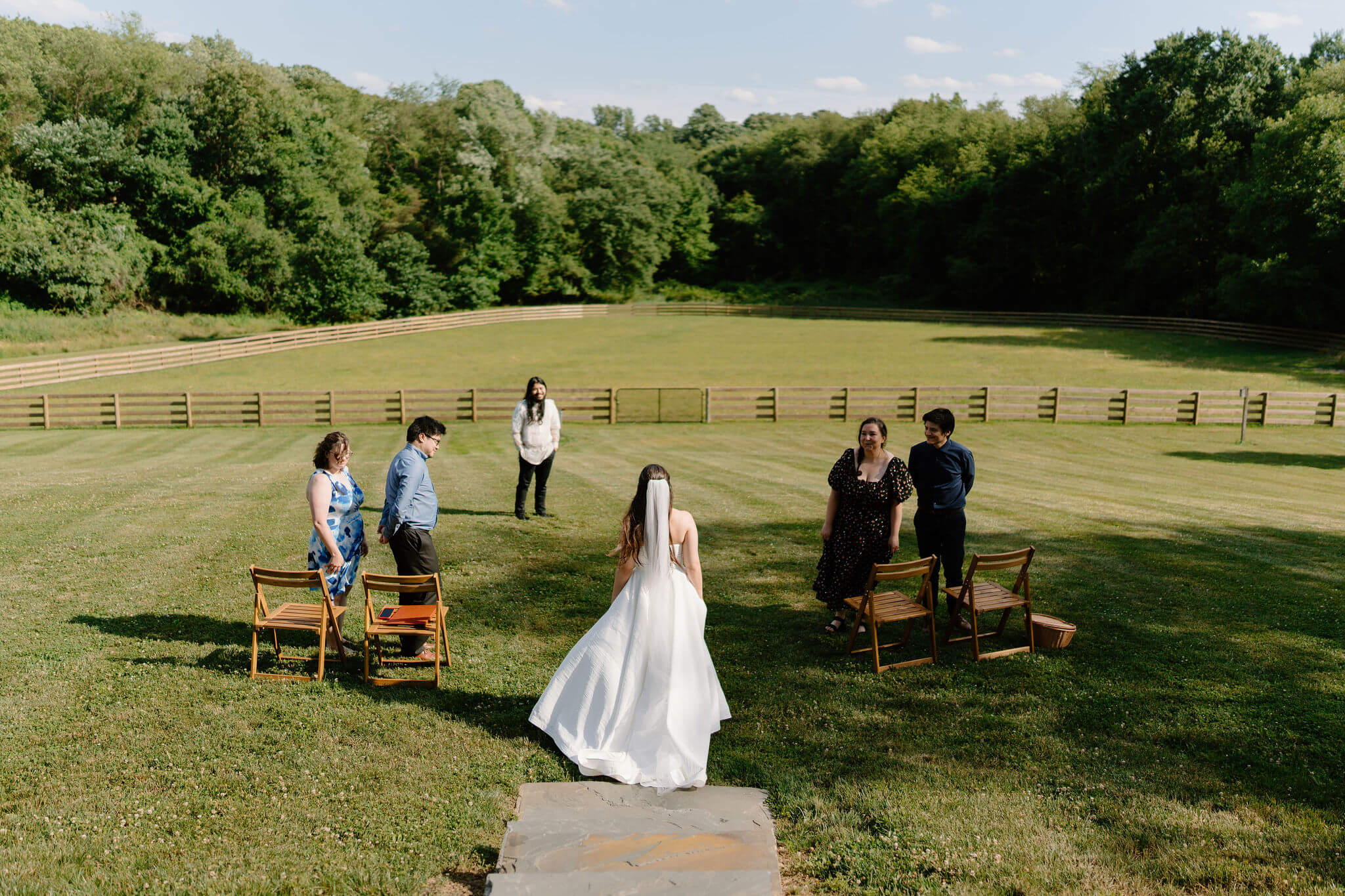 bride walking down a stone staircase into an open field to her elopement ceremony, with two wooden chairs on either side and two friends on either side smiling at her, and the groom (white shirt, black pants) smiling at her