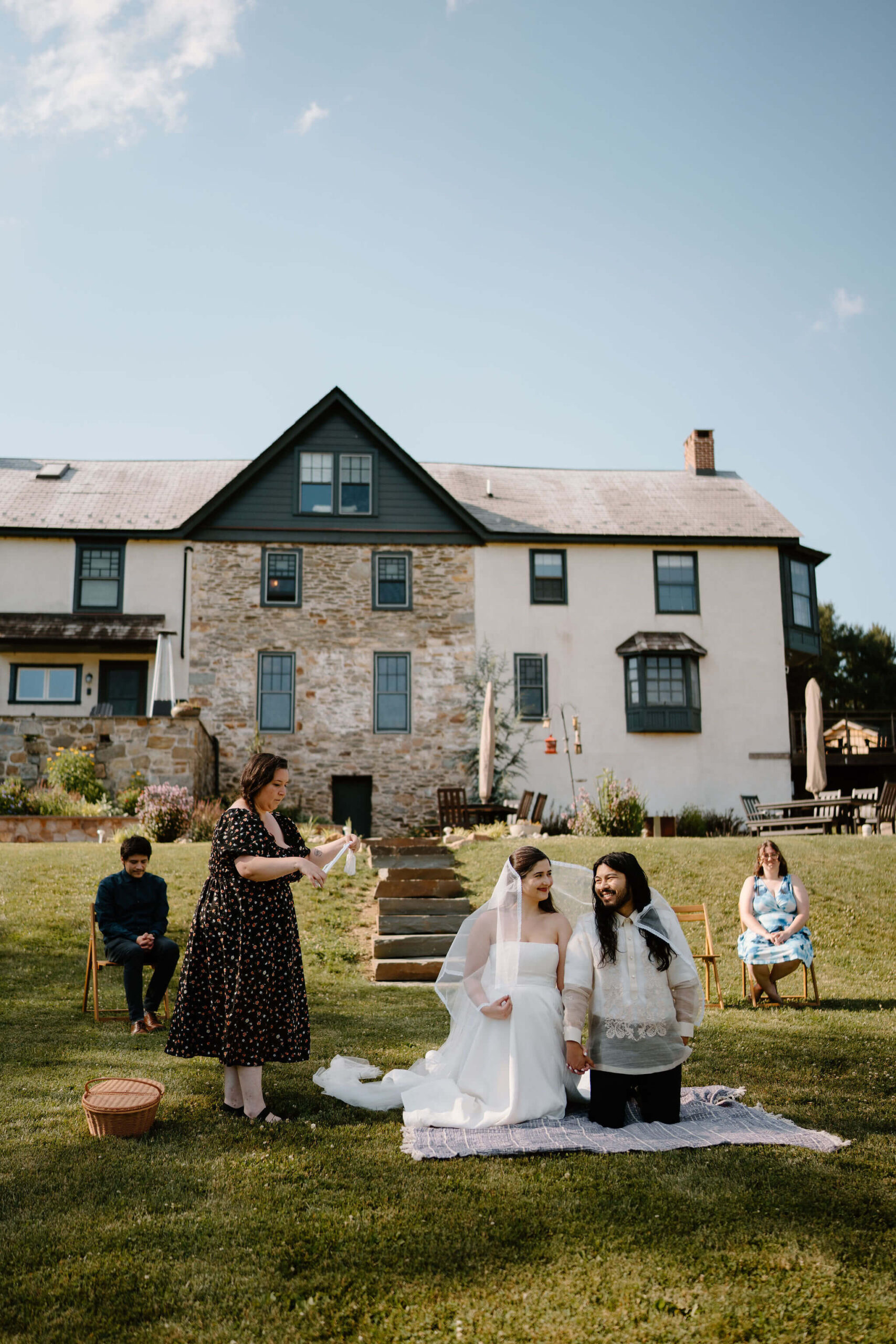 bride and groom kneeling on a blanket during their elopement ceremony, two guests seated behind them, with one guest standing next to them to perform a traditional Filipino lasso ceremony at a farmhouse in Pennsylvania