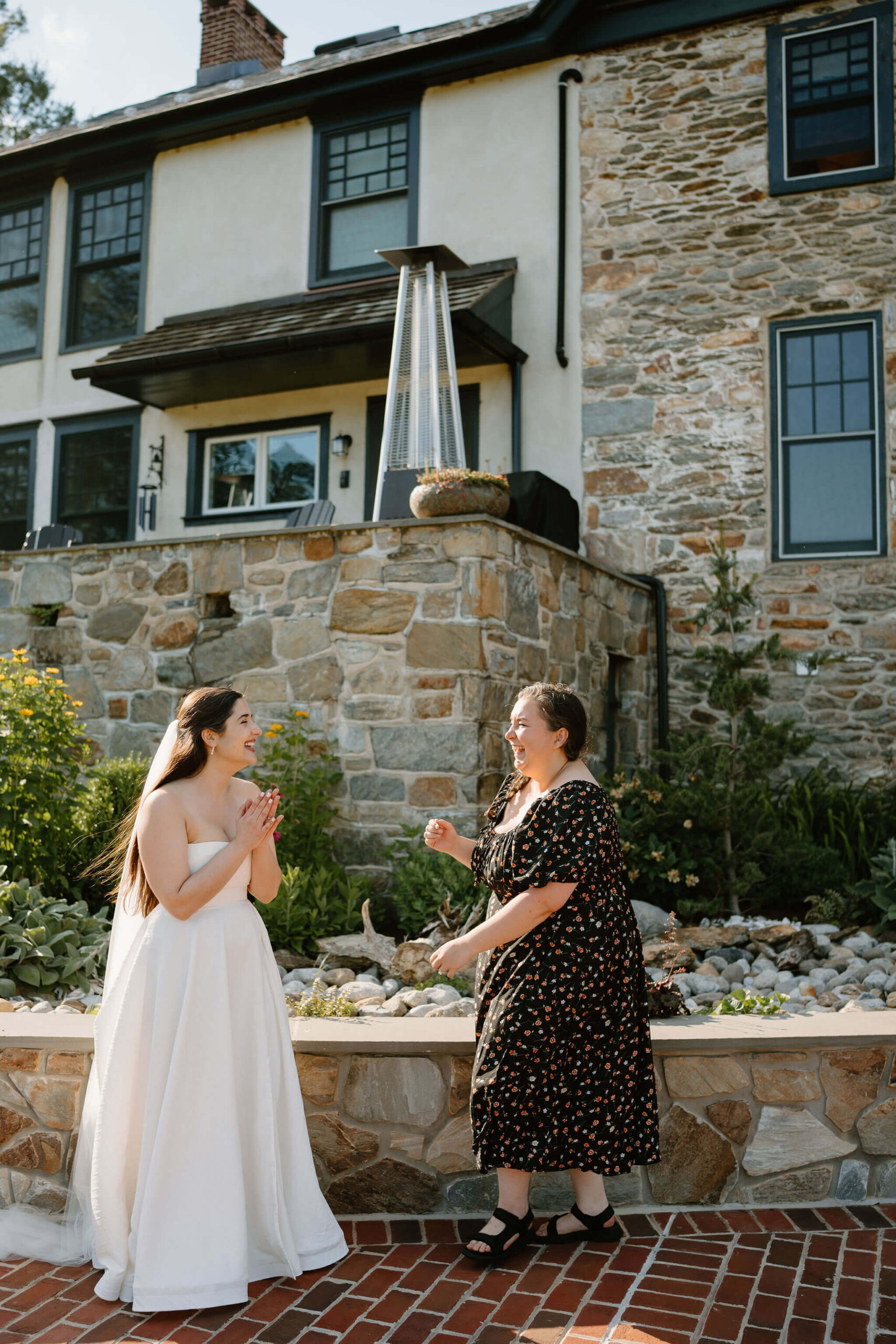 bride and her friend laughing loudly toward each other in front of a large stone farmhouse in Pennsylvania