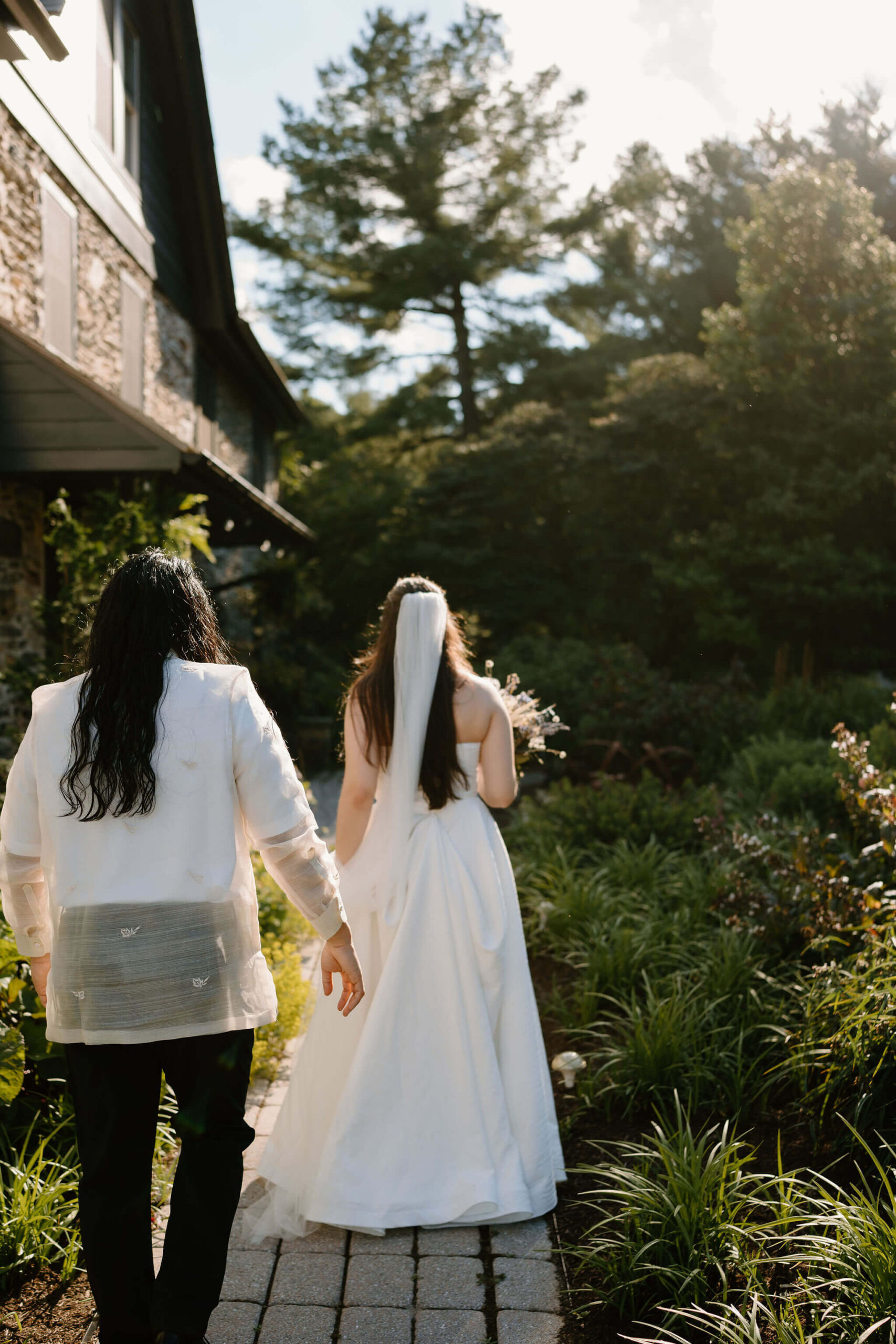 bride and groom walking up a stone garden path, away from the camera, sun backlighting them, next to a large stone farmhouse in Pennsylvania after eloping