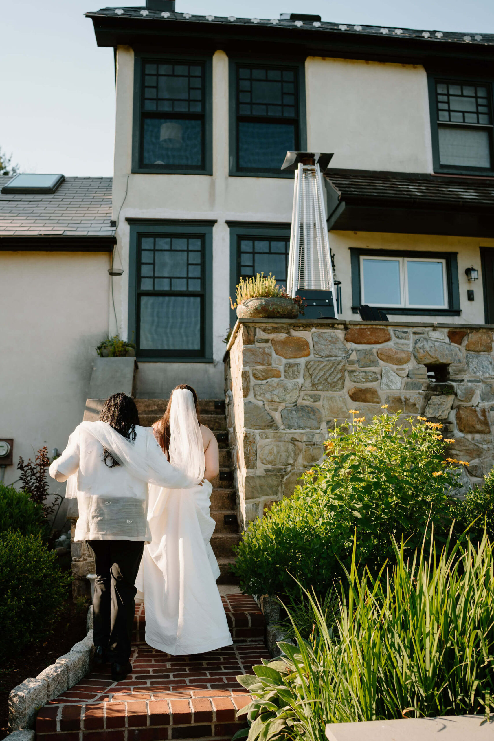 groom holding bride's veil over his shoulders while she walks up stairs to a stone patio at a Pennsylvania farmhouse, after eloping