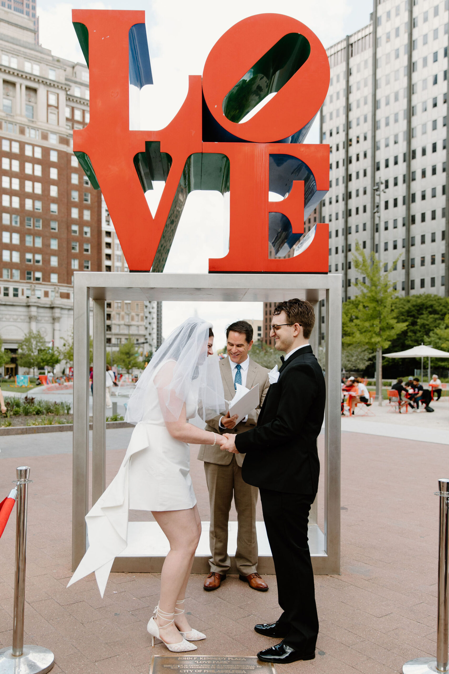 bride and groom laughing toward each other, standing with officiant in front of the Love Park sign during their elopement