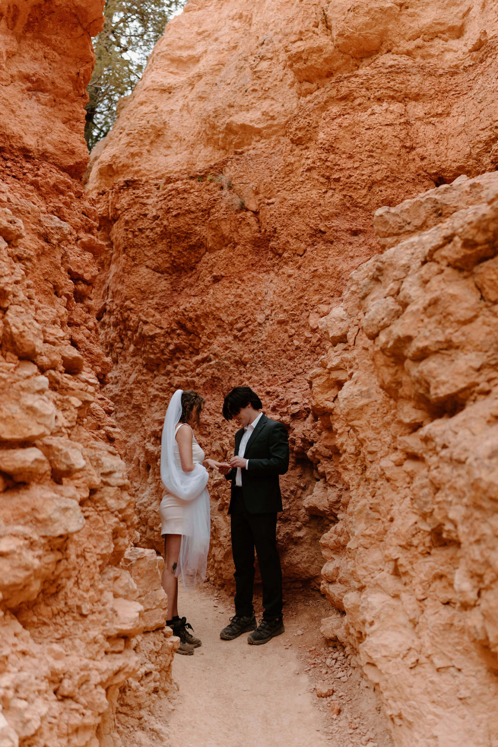 bride and groom on a narrow trail surrounded by tall red rocks, facing each other and exchanging wedding rings