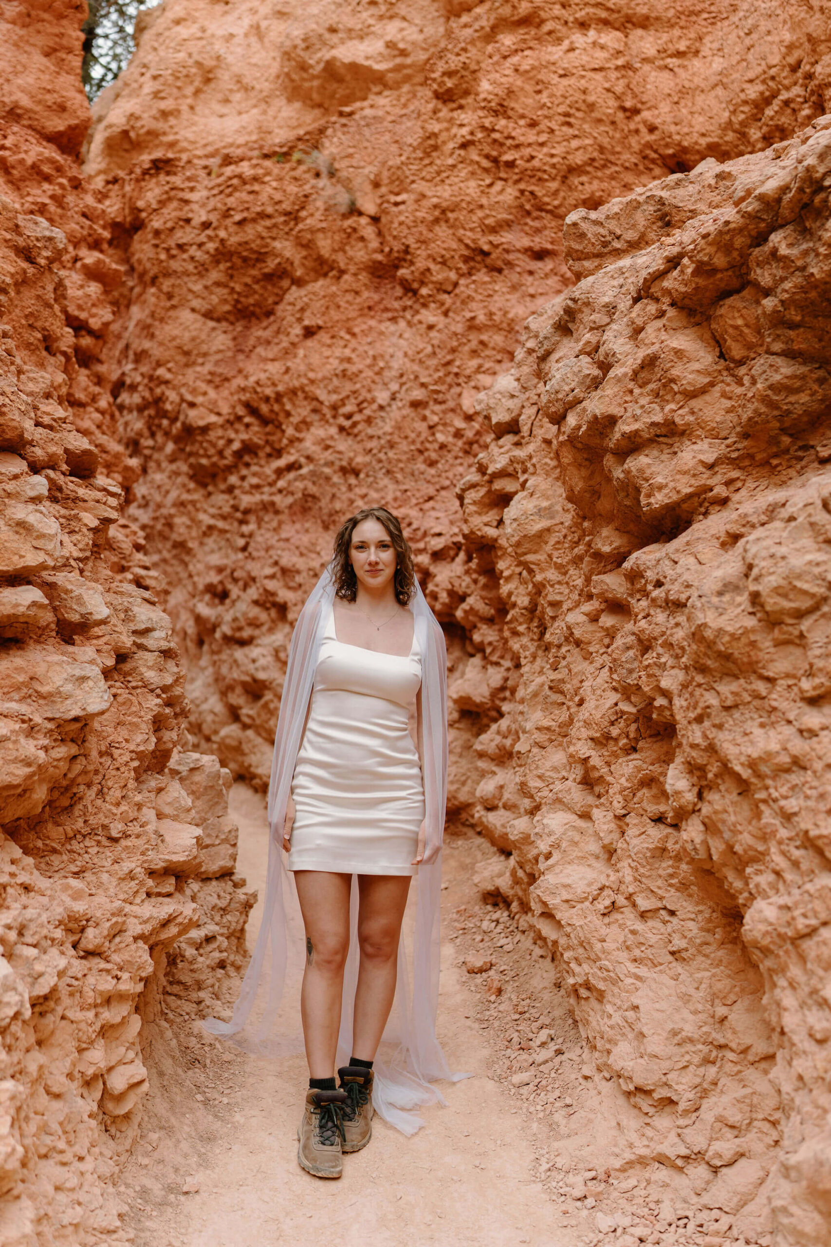bride wearing a short white wedding dress, a floor length veil, and hiking boots, smiling softly toward the camera on a narrow trail surrounded by tall red rocks
