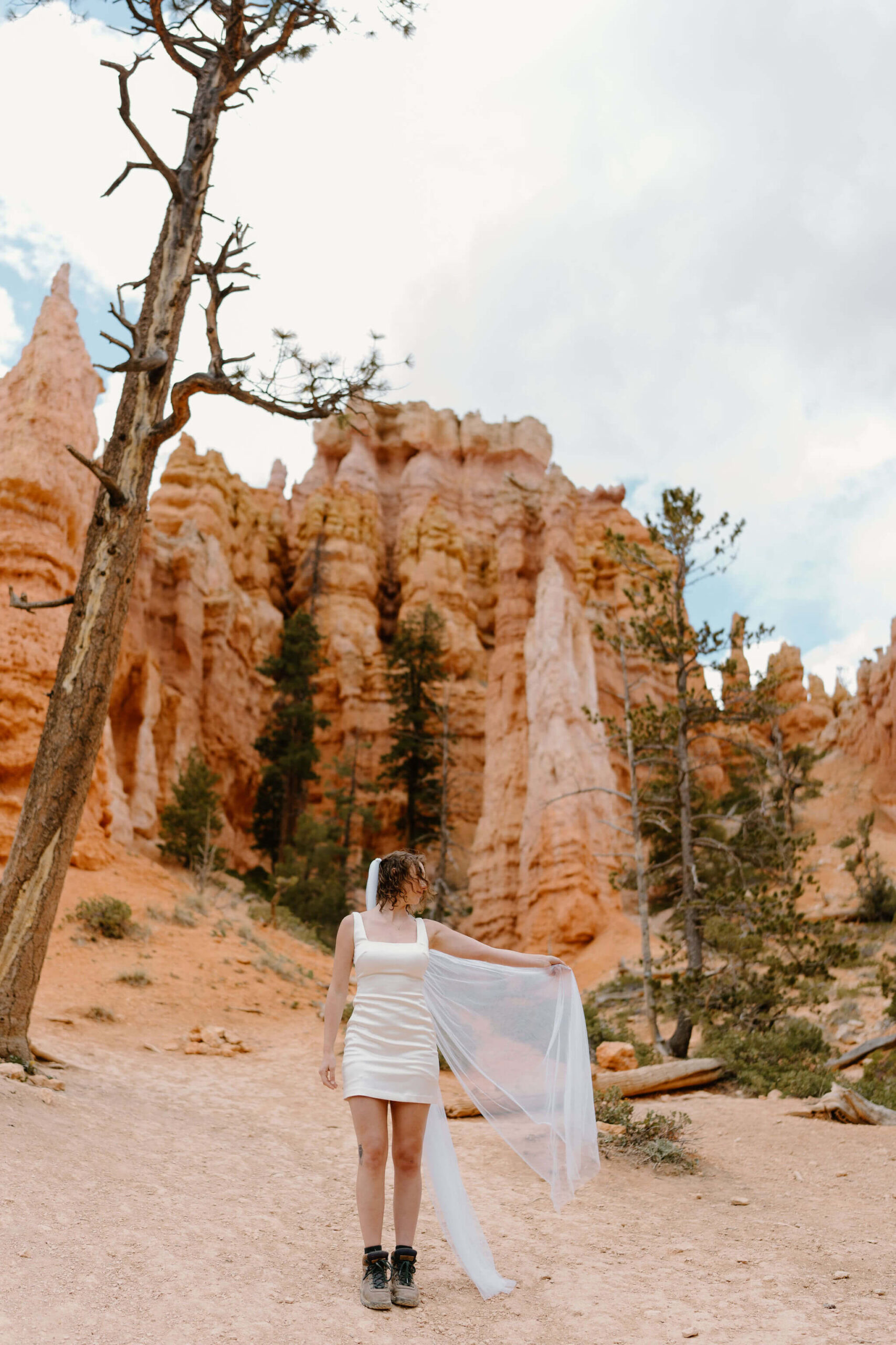 bride with a long white dress and long veil facing away from the camera, and looking over her shoulder off camera, holding flowers down at her side, with lots of trees and lush green bushes behind her