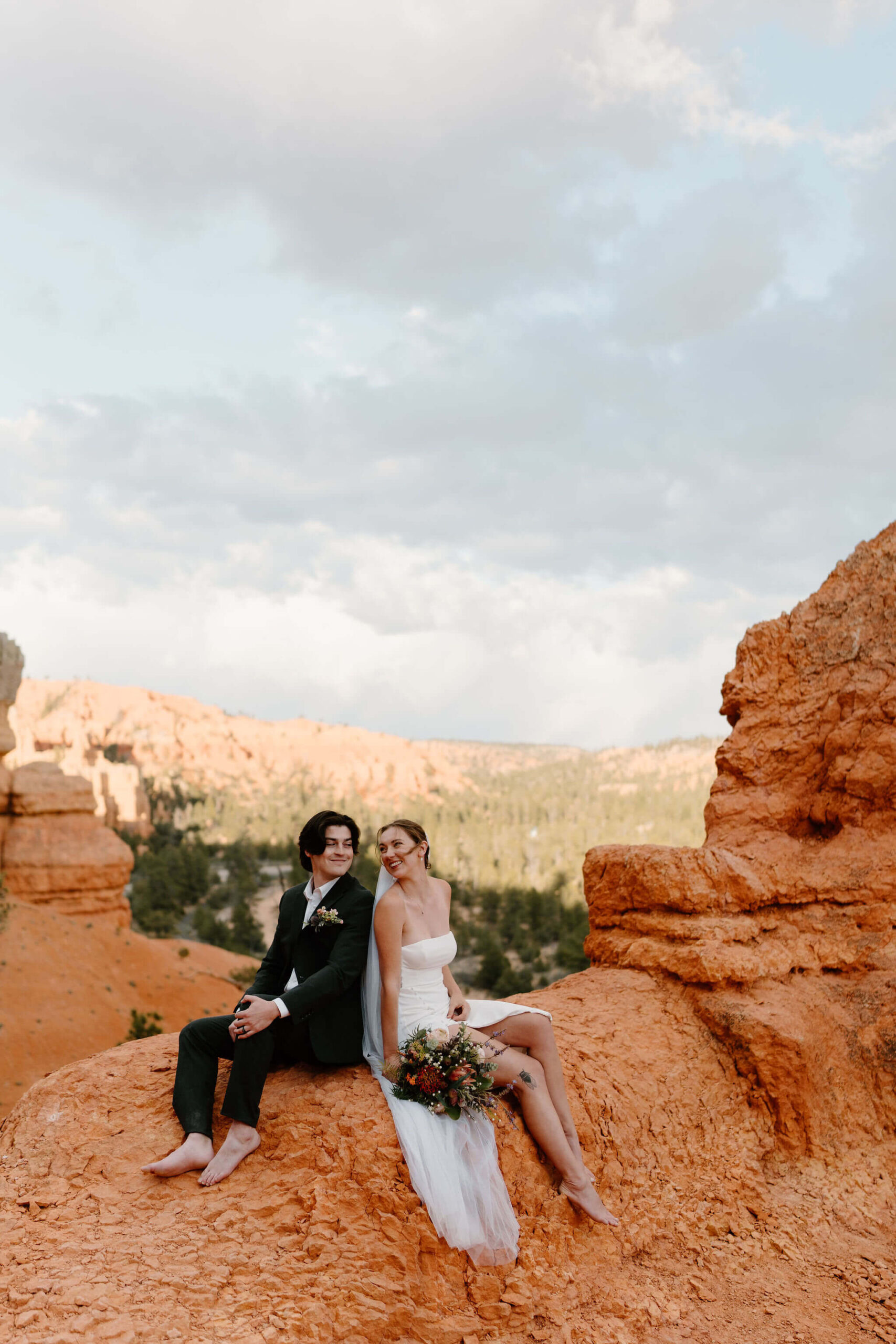 bride (white strapless gown, long brown hair, floor length veil) holding groom's face and kissing him on the cheek (white embroidered shirt, black pants, long brown hair) in front of a lush green garden with trees