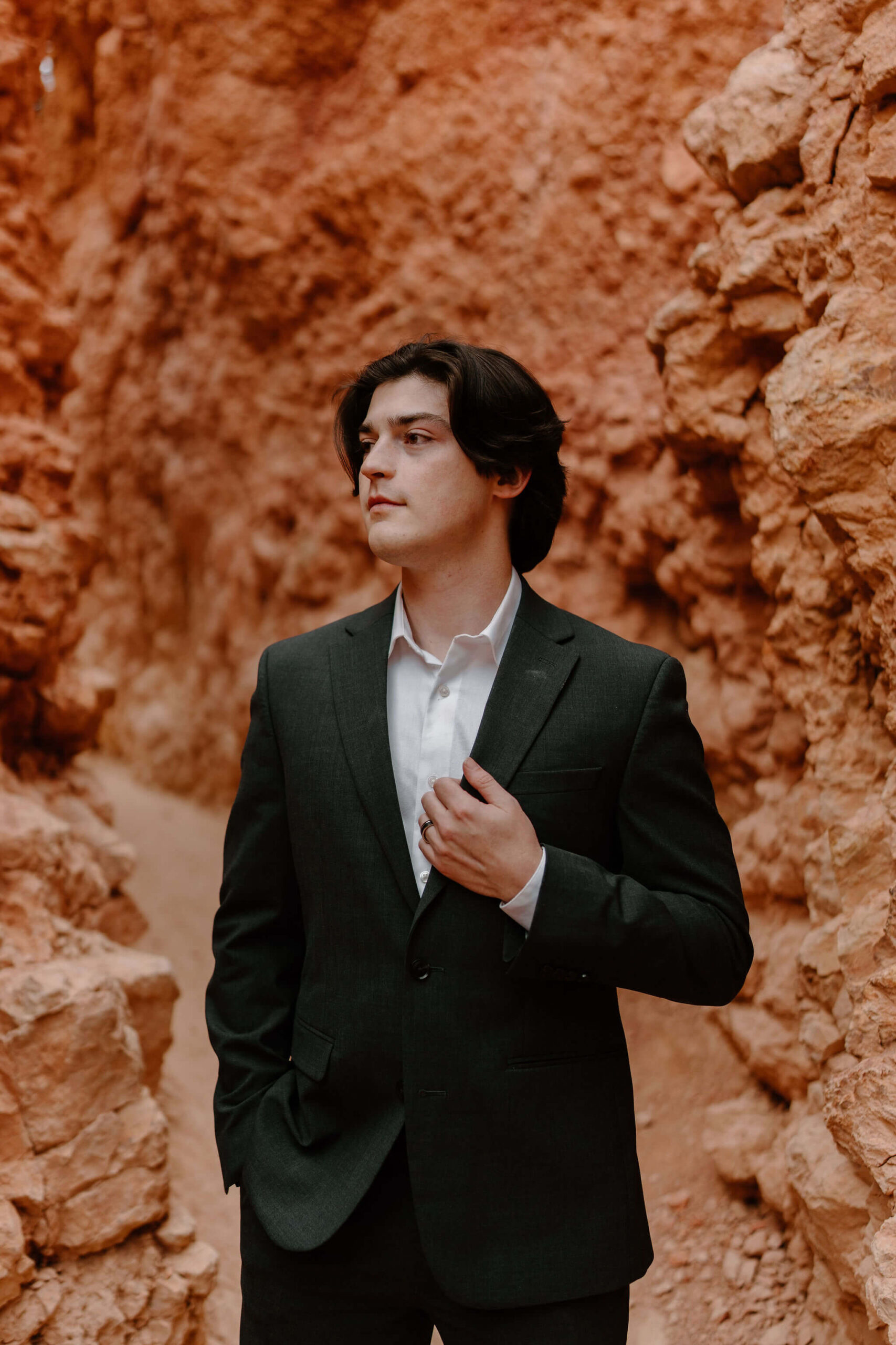 groom on a narrow trail surrounded by tall red rocks, holding his lapel with his left hand and looking off camera