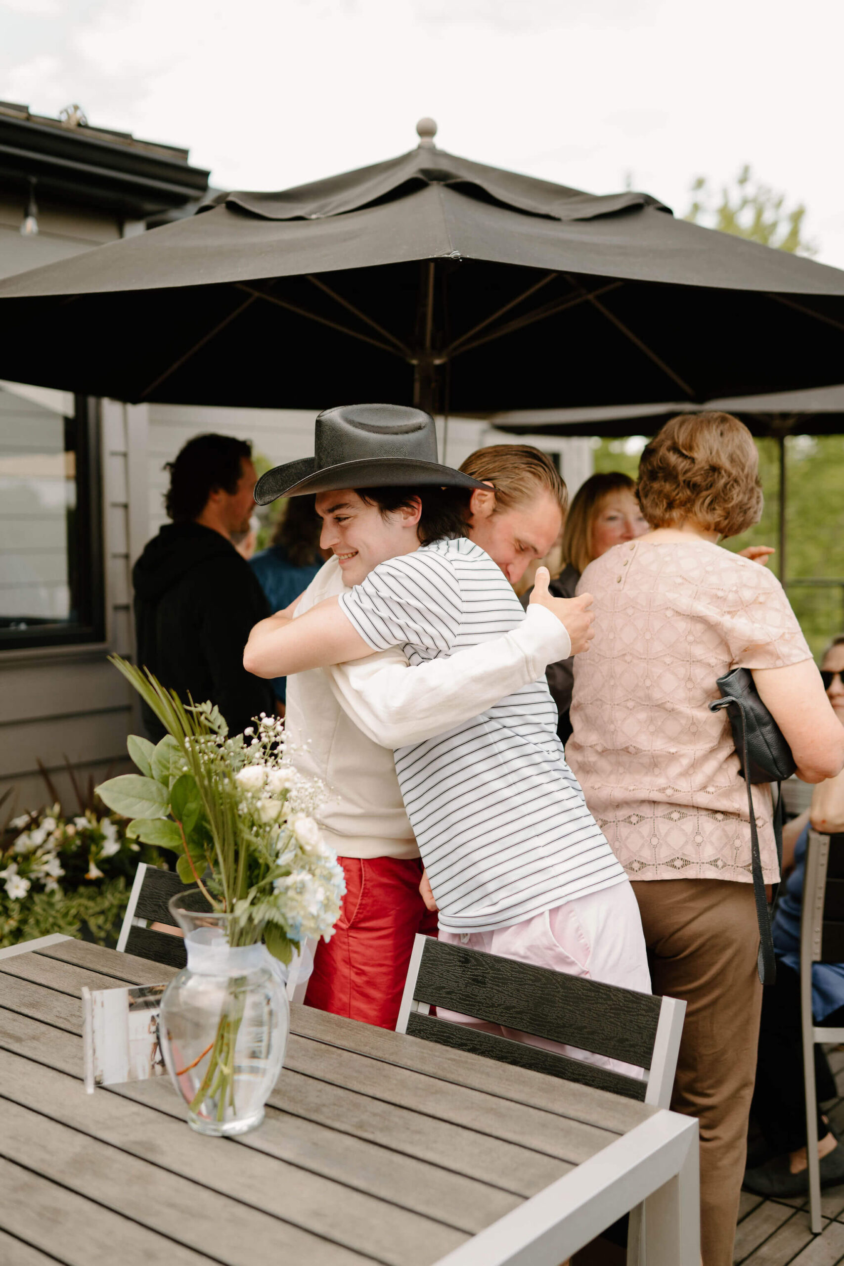 groom (striped polo shirt, black cowboy hat) hugging a guest and smiling