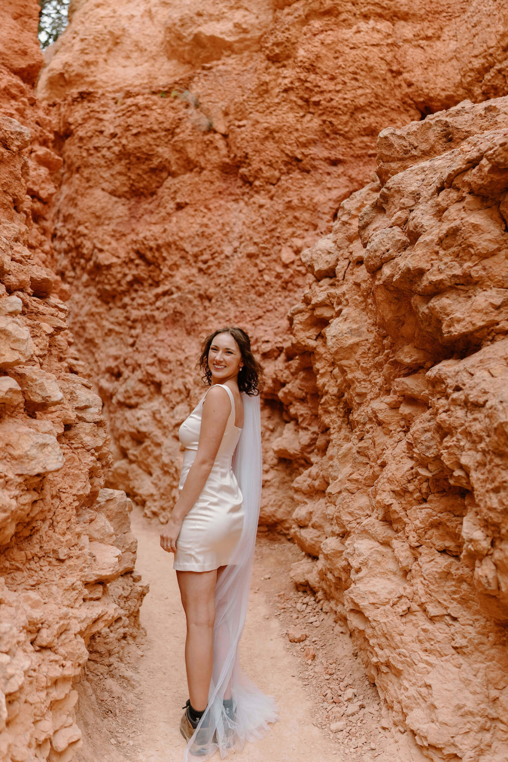 bride on a narrow trail surrounded by tall red rocks, looking over her shoulder toward the camera