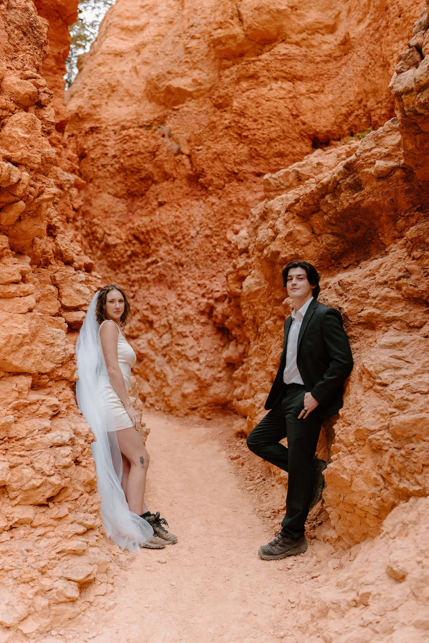 bride and groom leaning against rock formations on opposite sides of a hiking trial, smiling softly toward the camera
