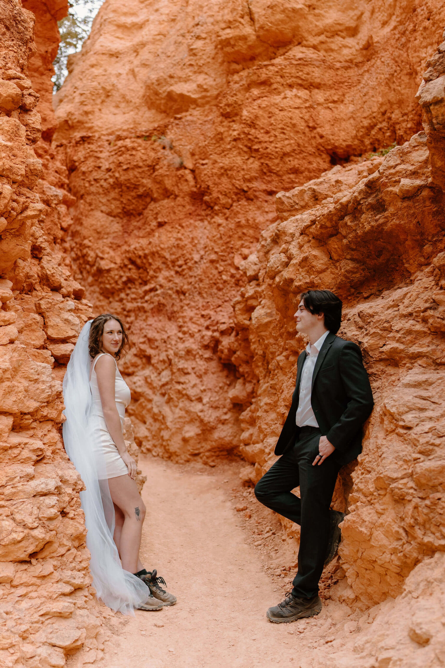 bride and groom leaning against rock formations on opposite sides of a hiking trial, smiling softly towards each other