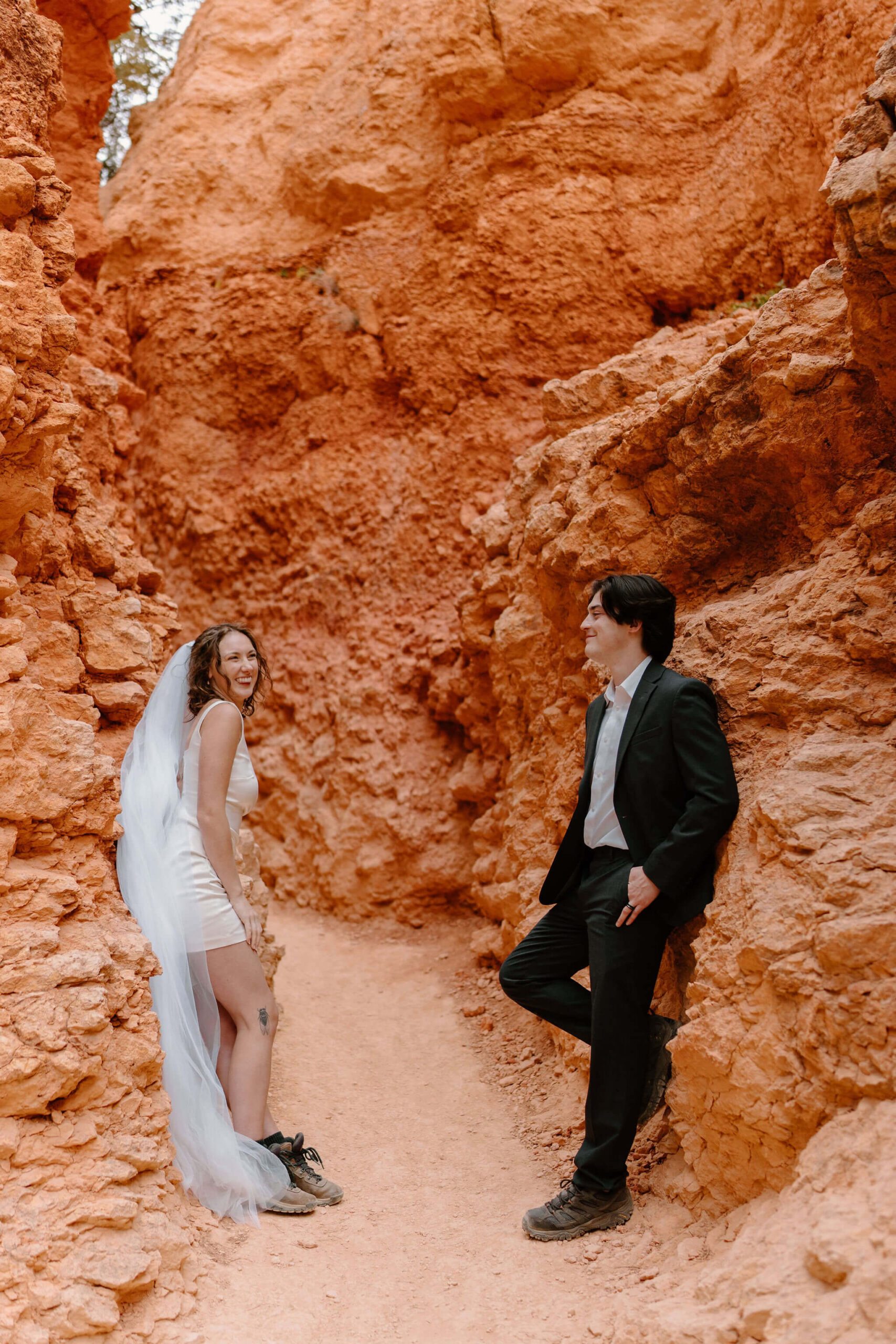 bride and groom leaning against rock formations on opposite sides of a hiking trial, laughing towards each other