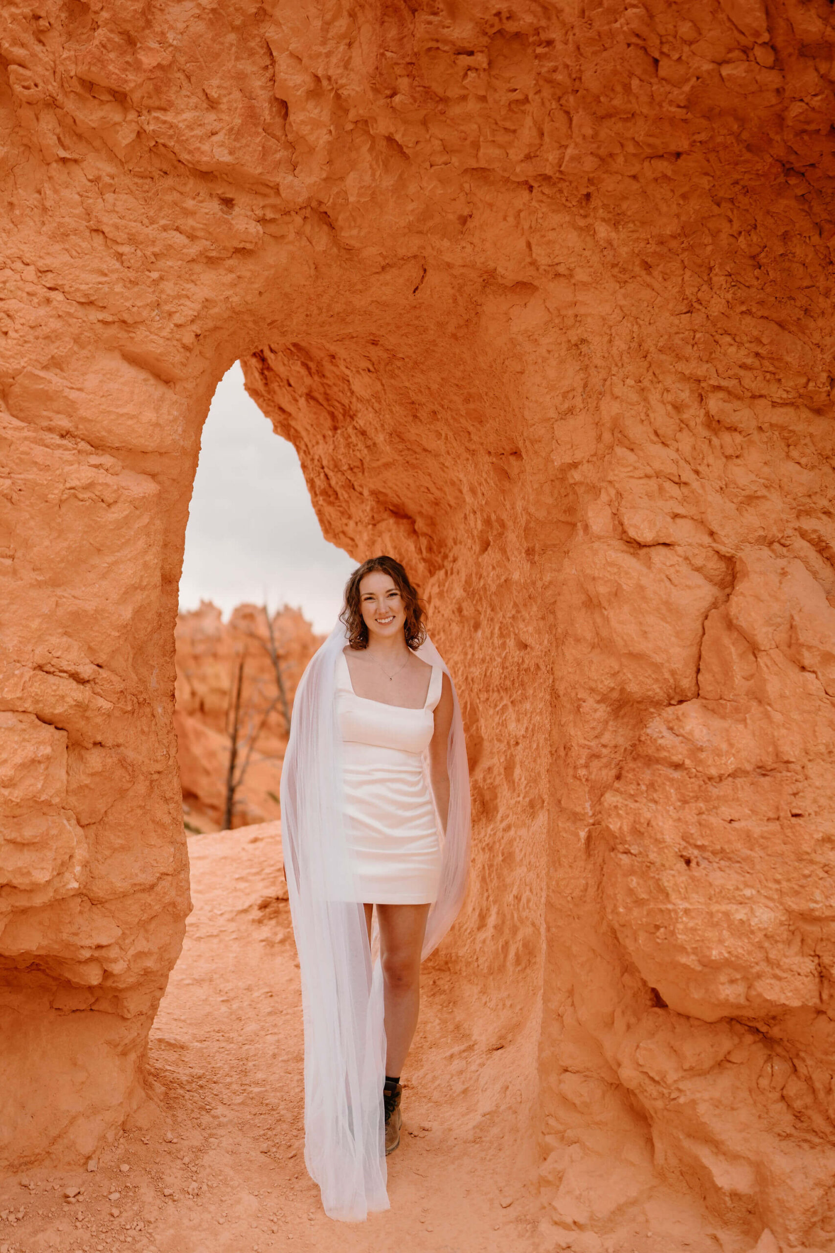 bride (short white wedding dress, floor length veil, hiking boots) standing in front of a small archway through a wall of red walks