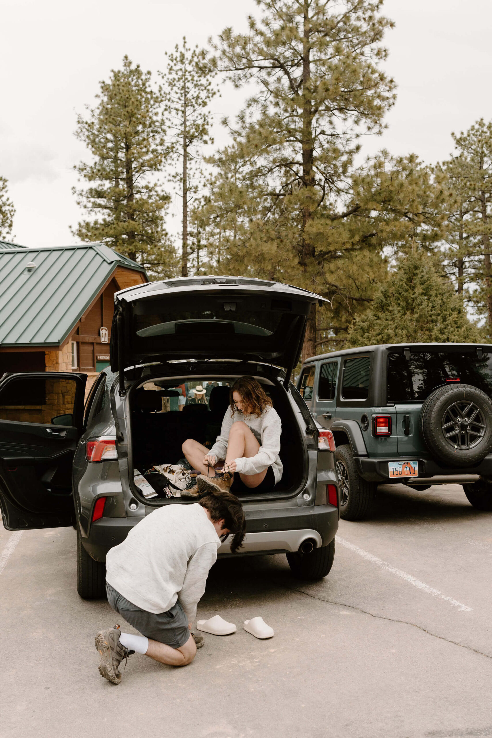 man kneeling behind an SUV tying his hiking boots, while a woman sits in the open trunk of the SUV lacing her hiking boots