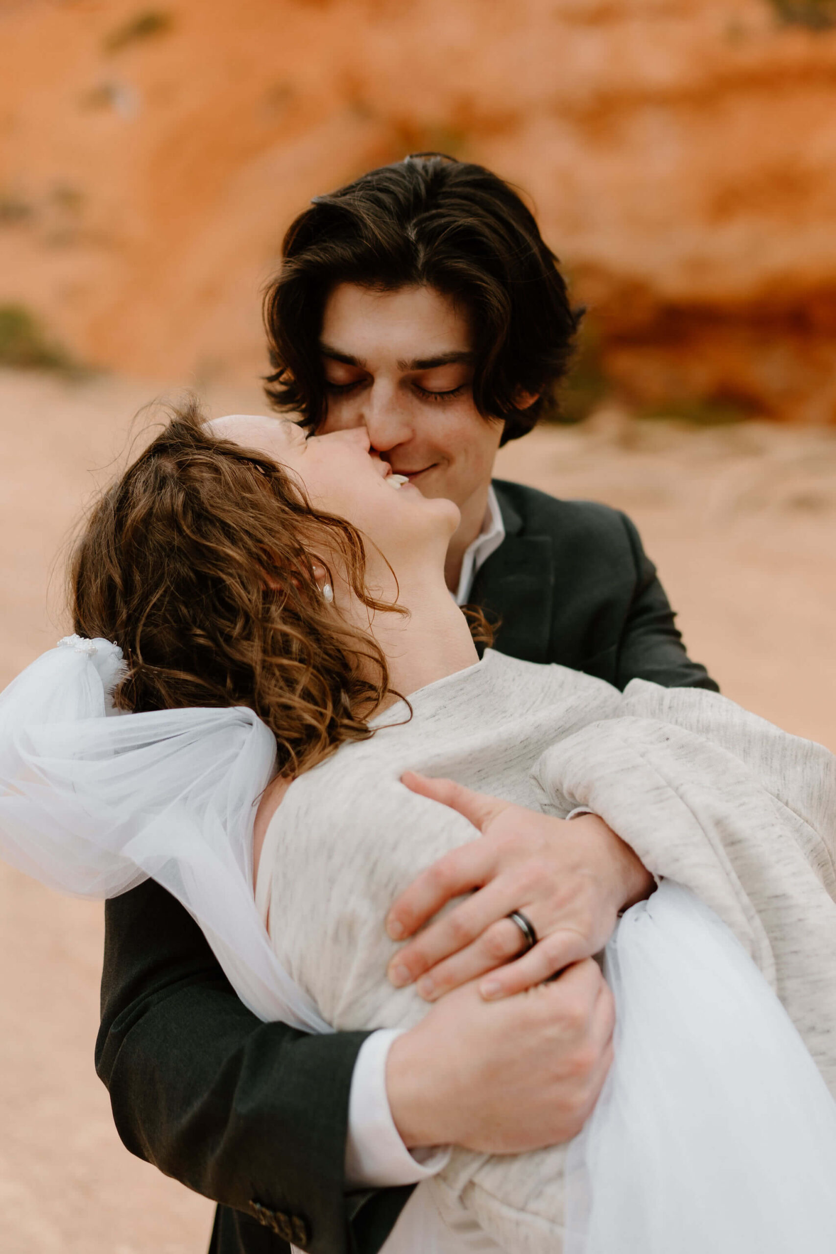 close up image of groom holding bride from behind, arms around her shoulders, smiling at each other, while she is wrapped up in a sweatshirt