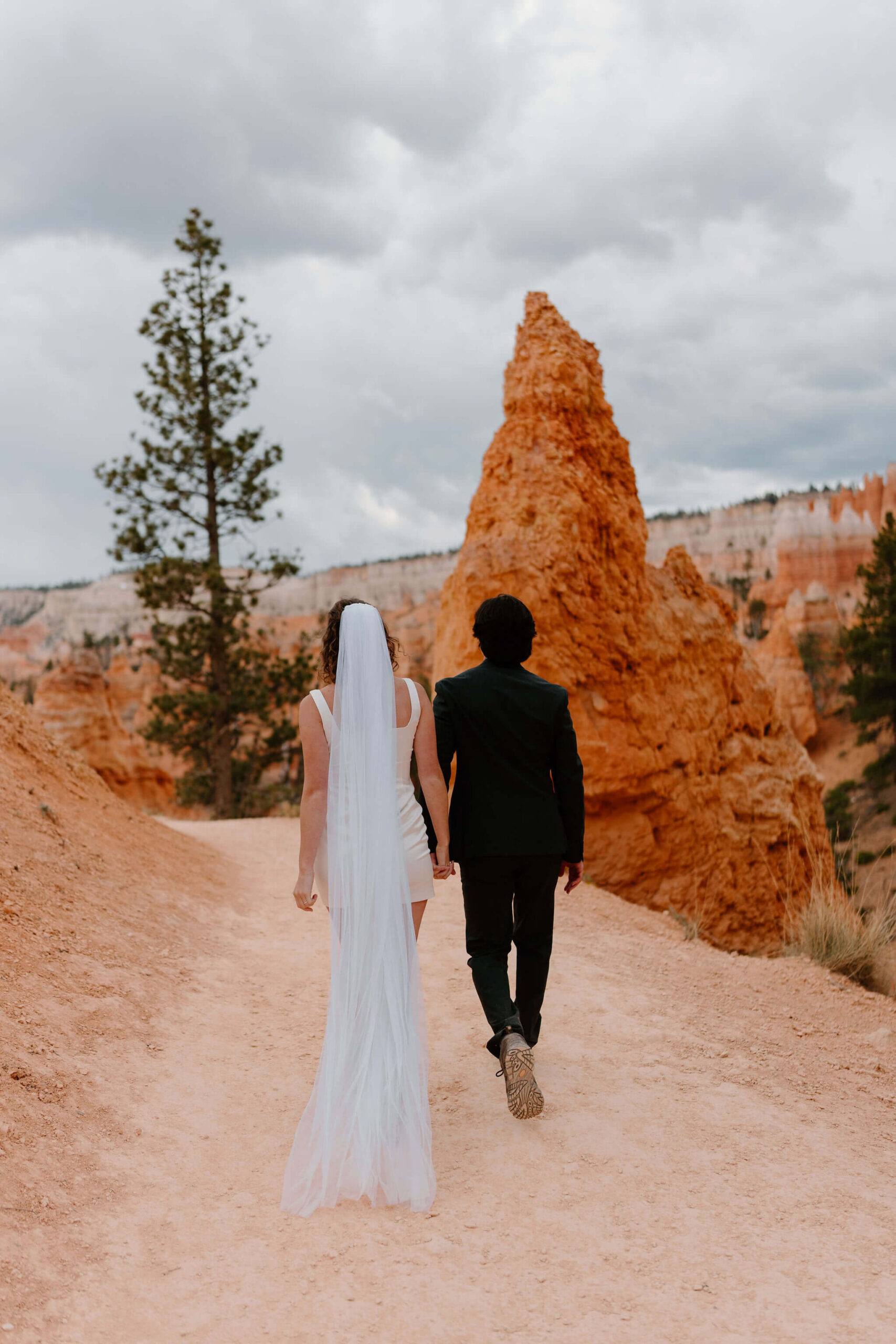 bride and groom walking down a trail, away from camera, through a canyon of red rocks