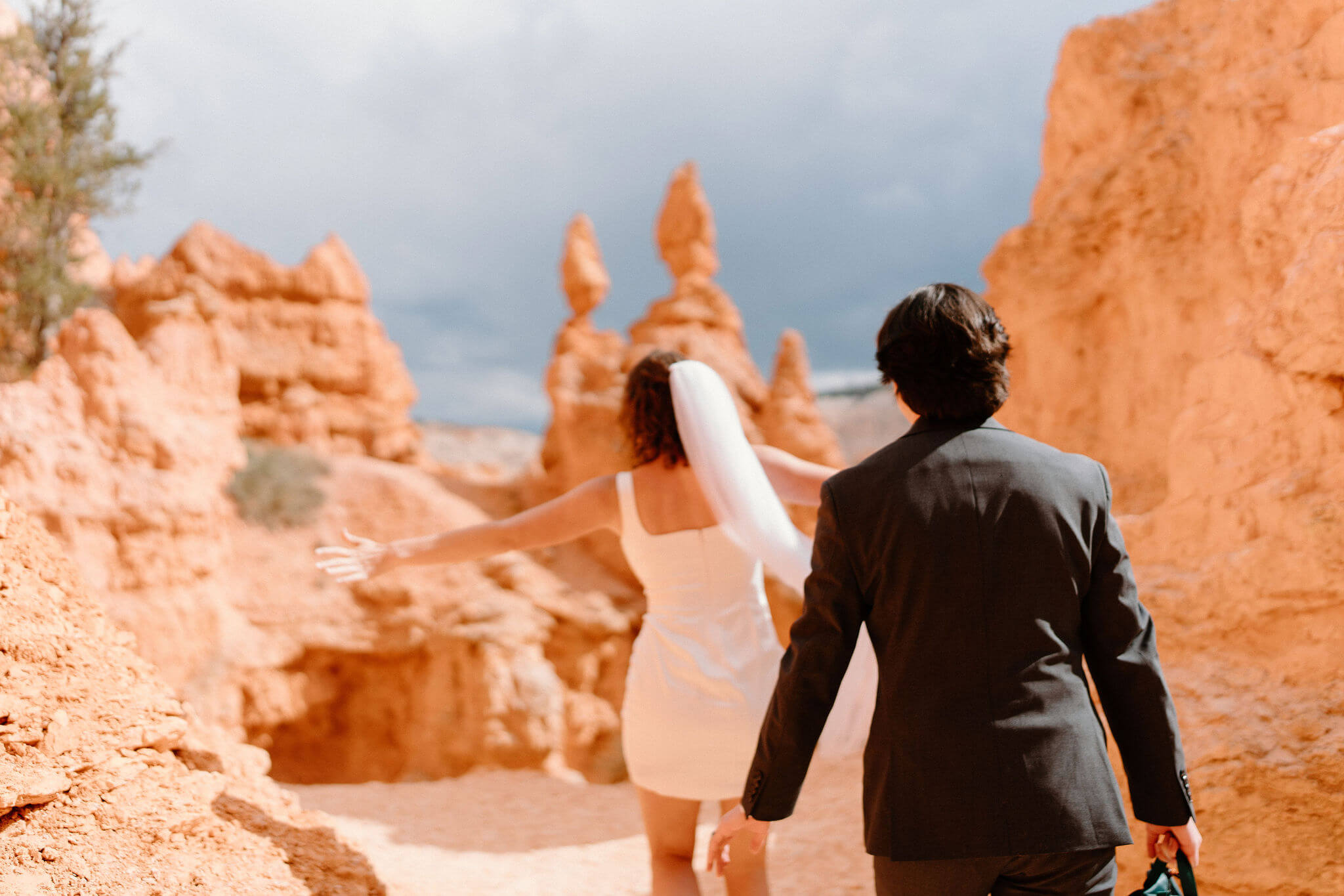 bride and groom walking through tall red hoodoos, with dark storm clouds in the distance