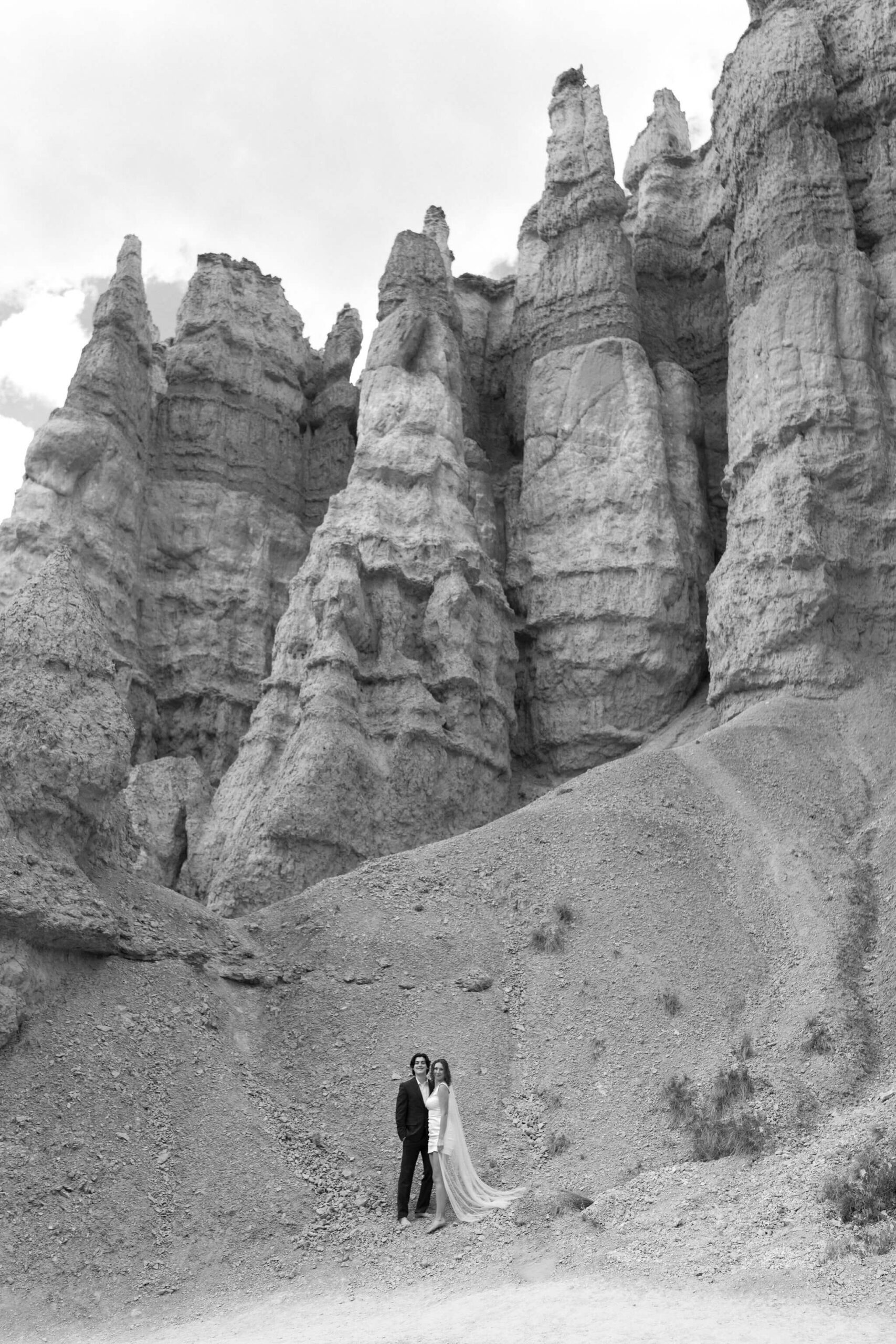 black and white image of bride and groom embracing with a large wall of hoodoos behind them