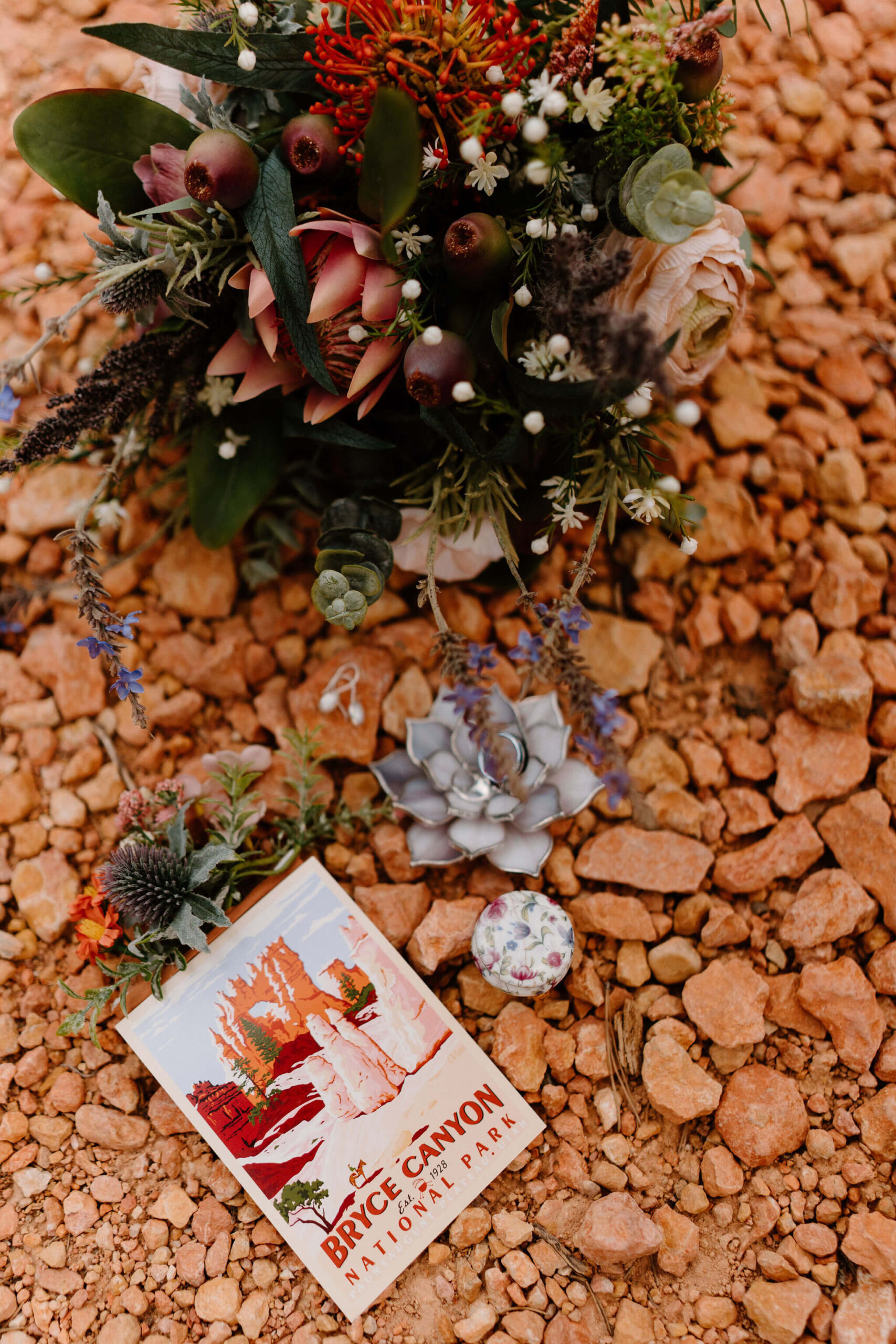 flat lay of a colorful bouquet of flowers, earrings, a lotus flower ring dish with two wedding bands, a pocket boutonniere, and a post card of Bryce Canyon National Park, against a background of small red rocks