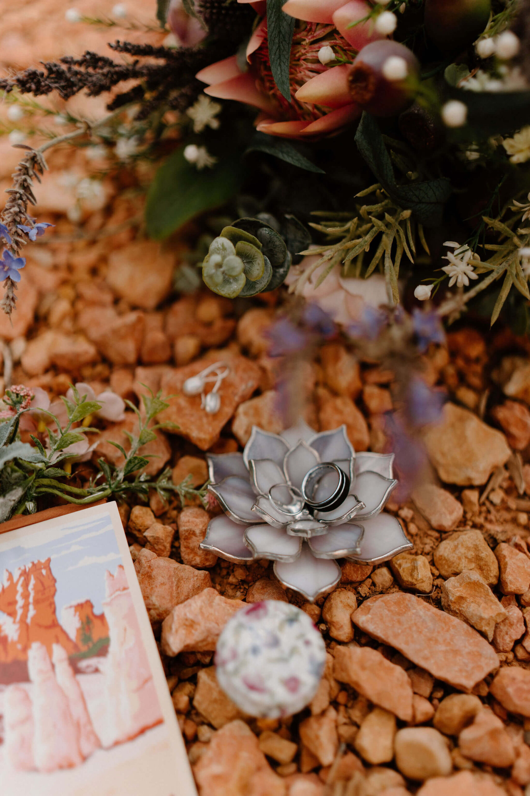 flat lay of a colorful bouquet of flowers, earrings, a lotus flower ring dish with two wedding bands, a pocket boutonniere, and a post card of Bryce Canyon National Park, against a background of small red rocks