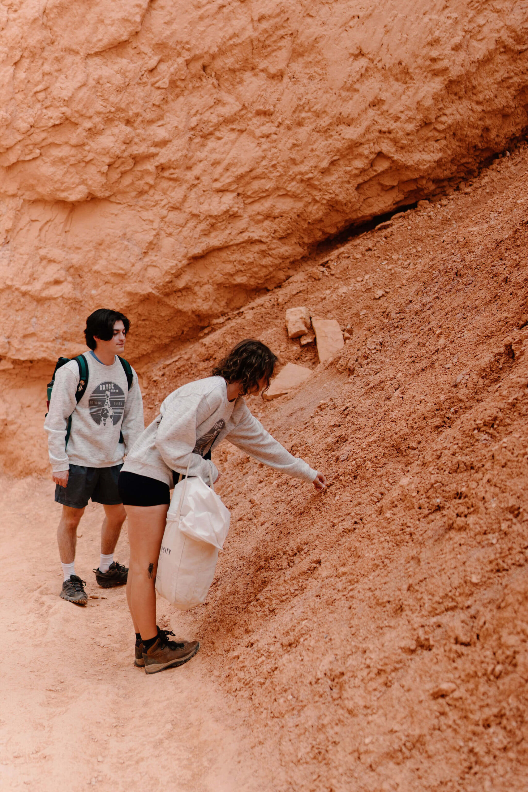 woman holding a garment bag and pointing at red soil, while a man watches behind her at Bryce Canyon National Park.