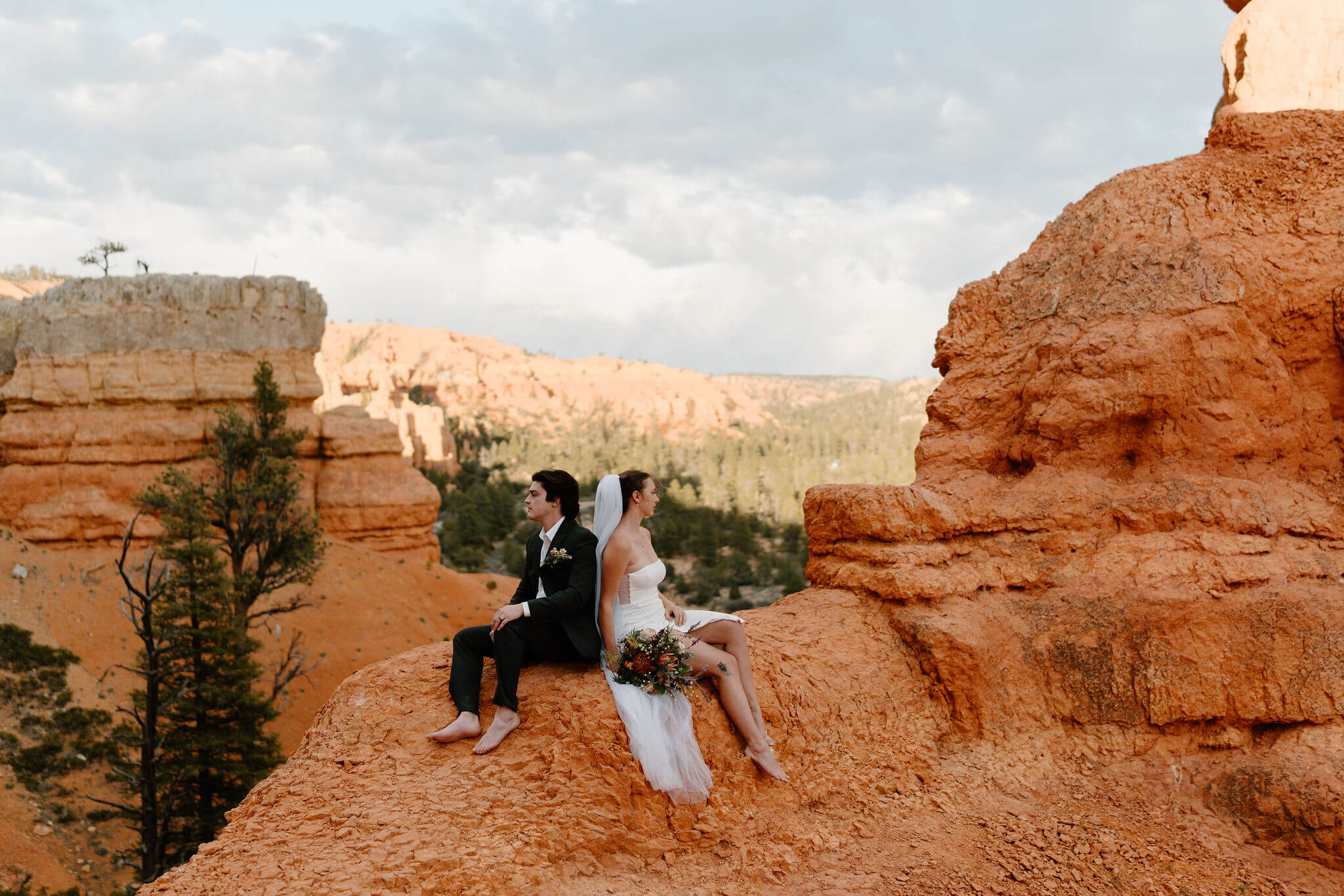 bride and groom sitting on a red rock overlook, back to back, with red cliffs all around them at the Red Canyon Trailhead